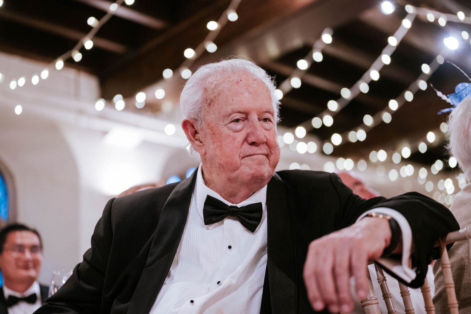 An elderly gentleman in formal black tie attire sits at what appears to be an elegant event.