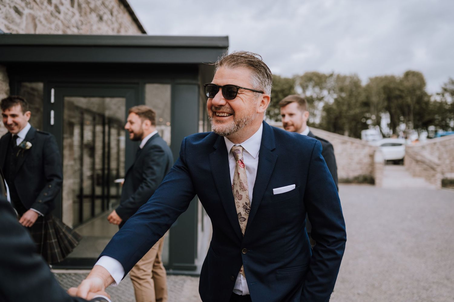 Stylishly dressed guest in blue suit and sunglasses smiling at outdoor event.