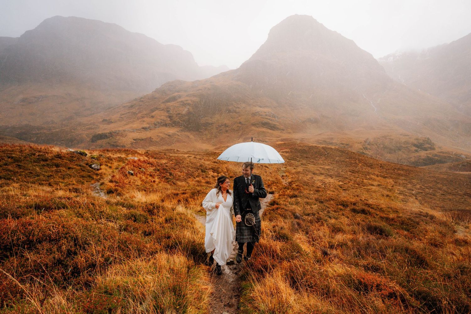 Couple holding a white umbrella against a backdrop of misty Scottish highlands with golden autumn colors.