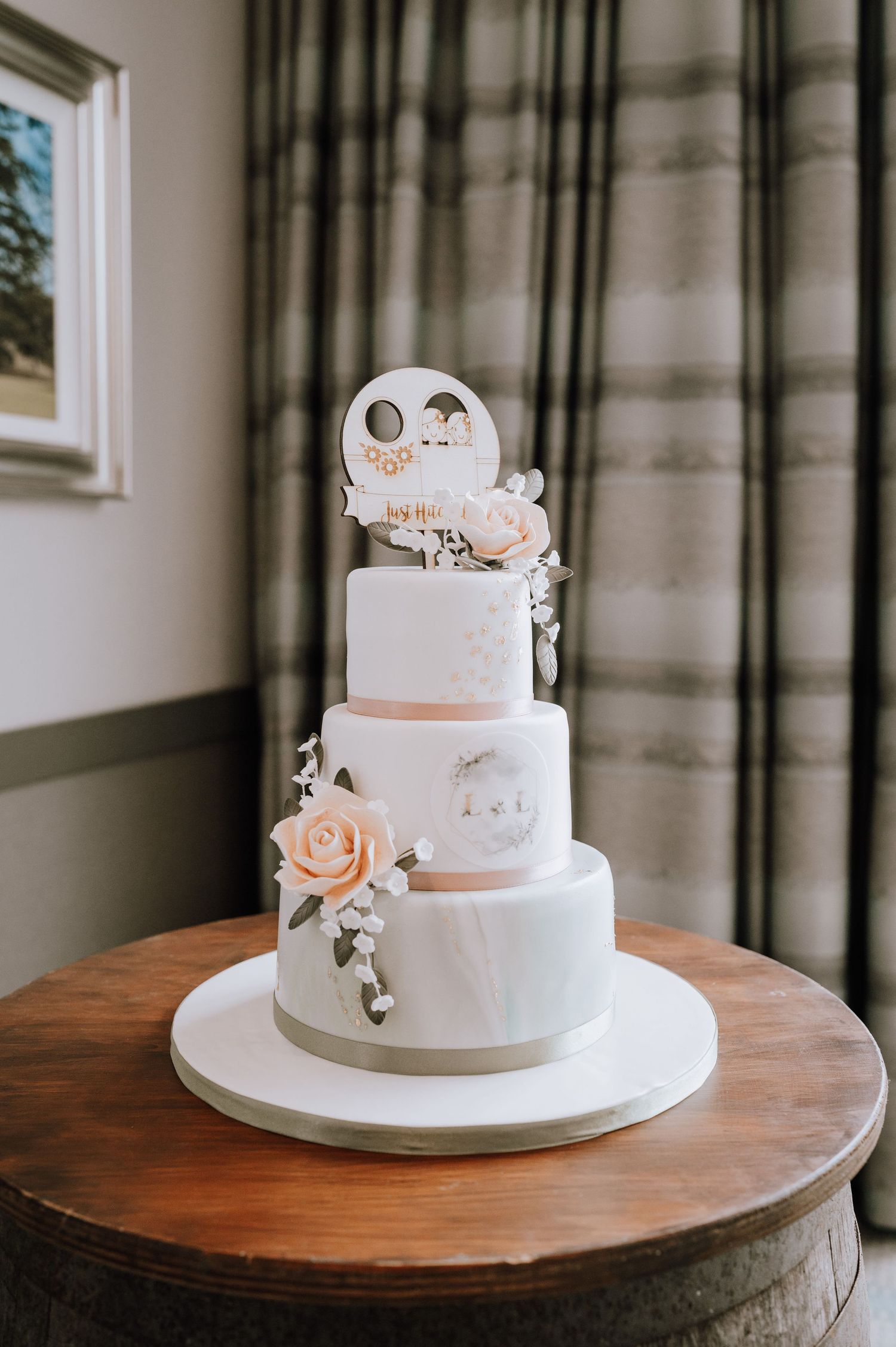 Elegant three-tier white wedding cake with peach roses and custom topper on wooden table.