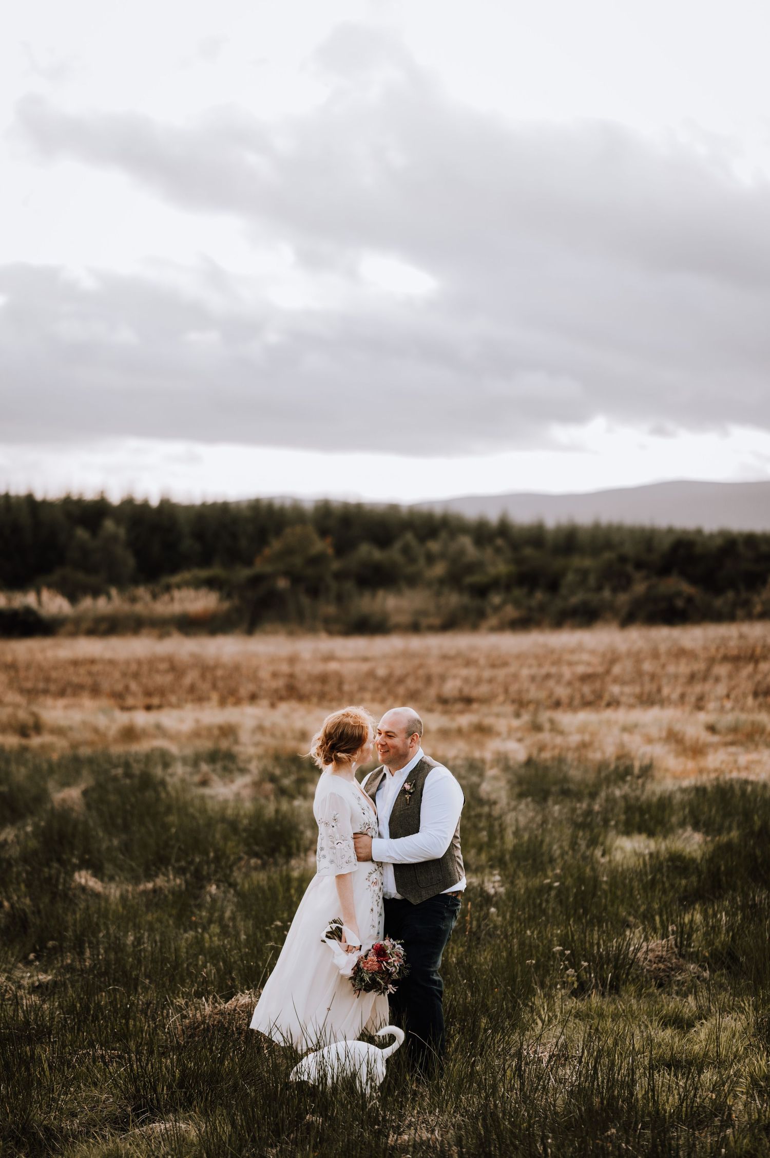 A couple shares an intimate moment in a scenic field under dramatic cloudy skies.
