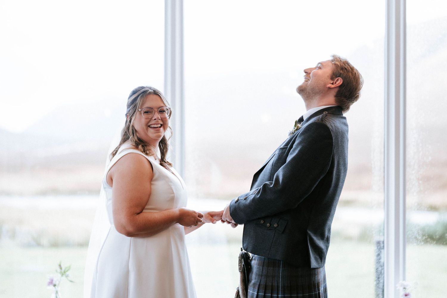 A joyful couple shares a laugh during their wedding ceremony in front of bright windows.