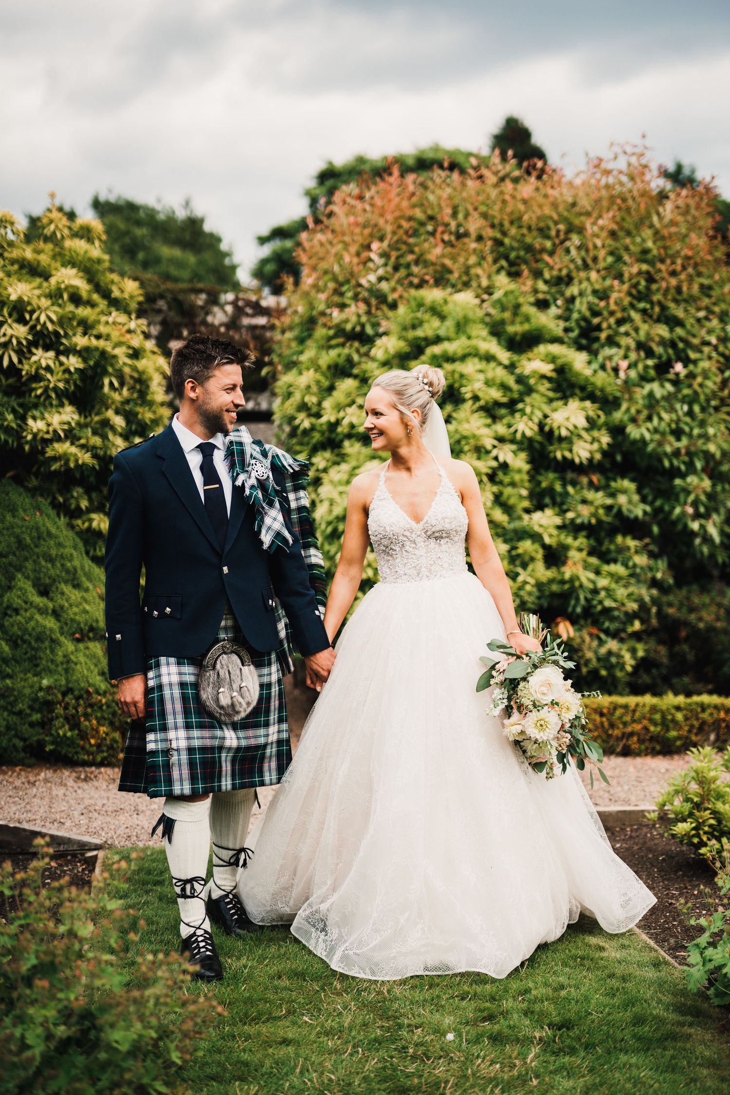 Bride in white ballgown and groom in traditional Scottish kilt walking together in a garden setting.