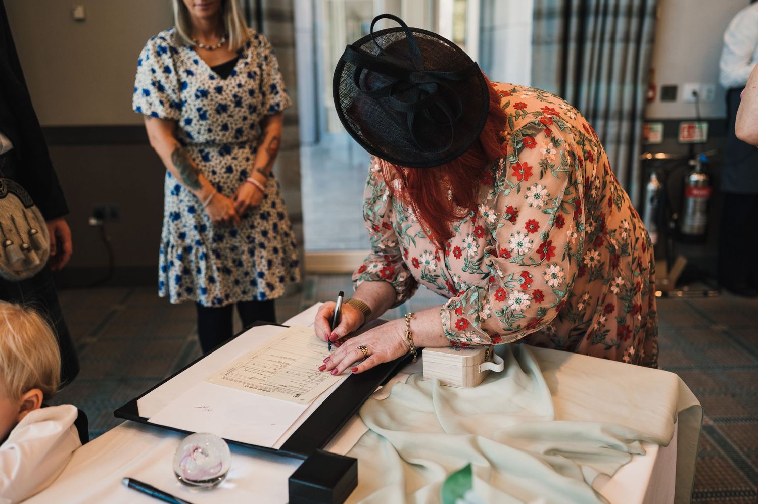 Wedding guests in floral dresses signing documents at an elegant indoor ceremony.
