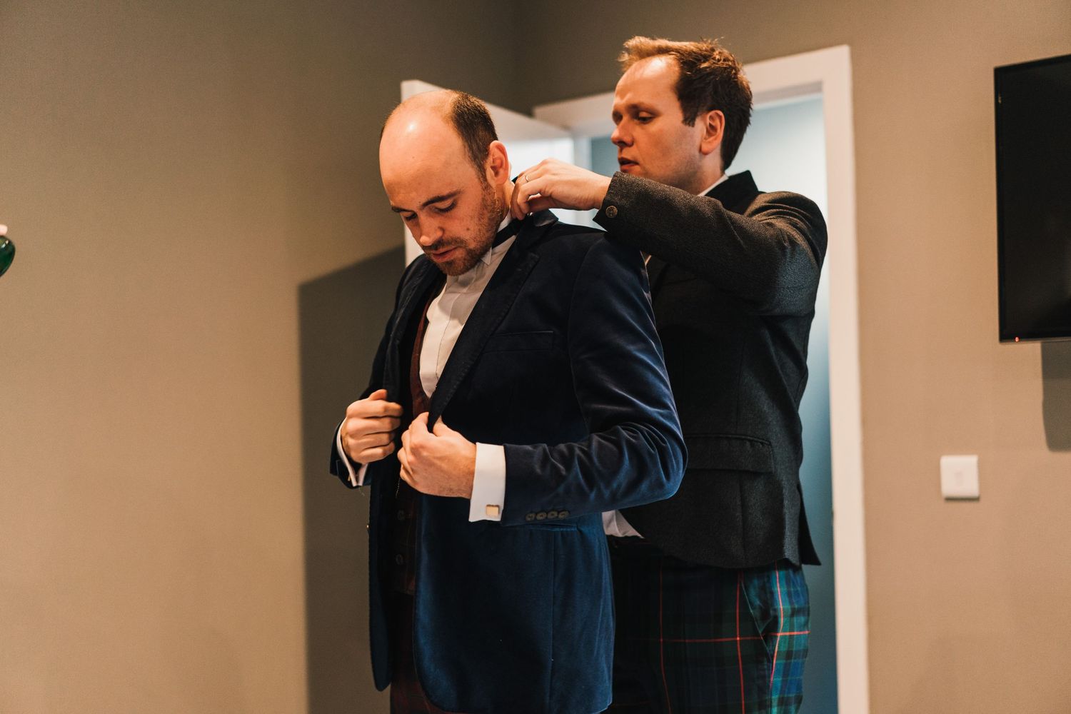 One person helps another adjust their formal navy jacket during wedding preparations.