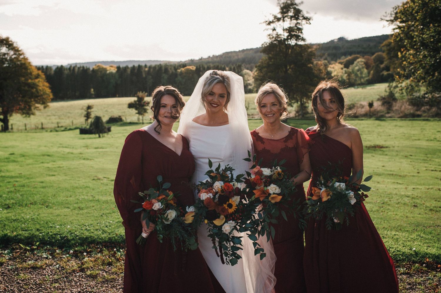Bridal party in burgundy dresses holding bouquets pose together in a scenic outdoor setting.