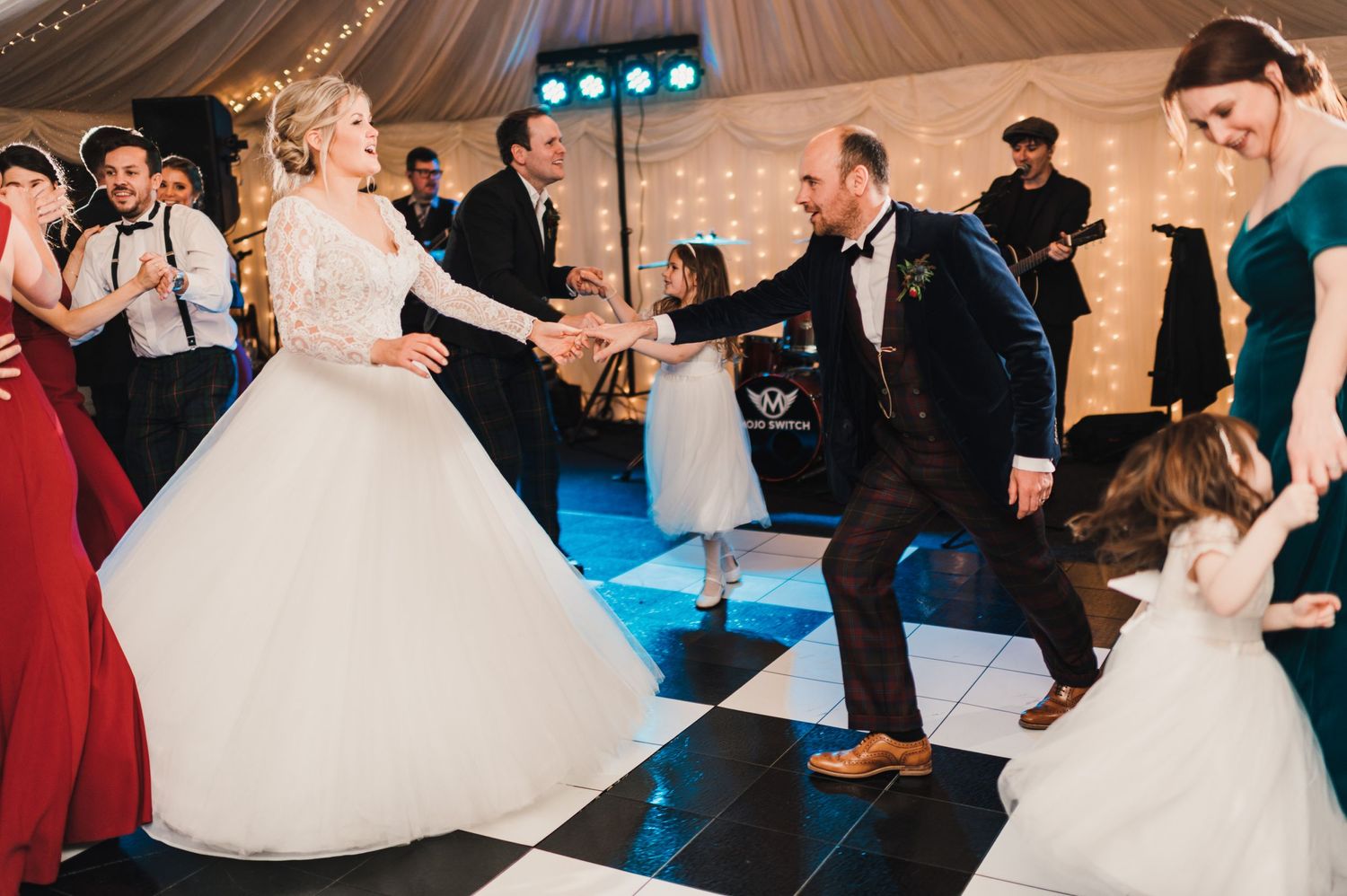 Wedding couple dances with guests on a black and white checkered dance floor beneath elegant draped tent ceiling.