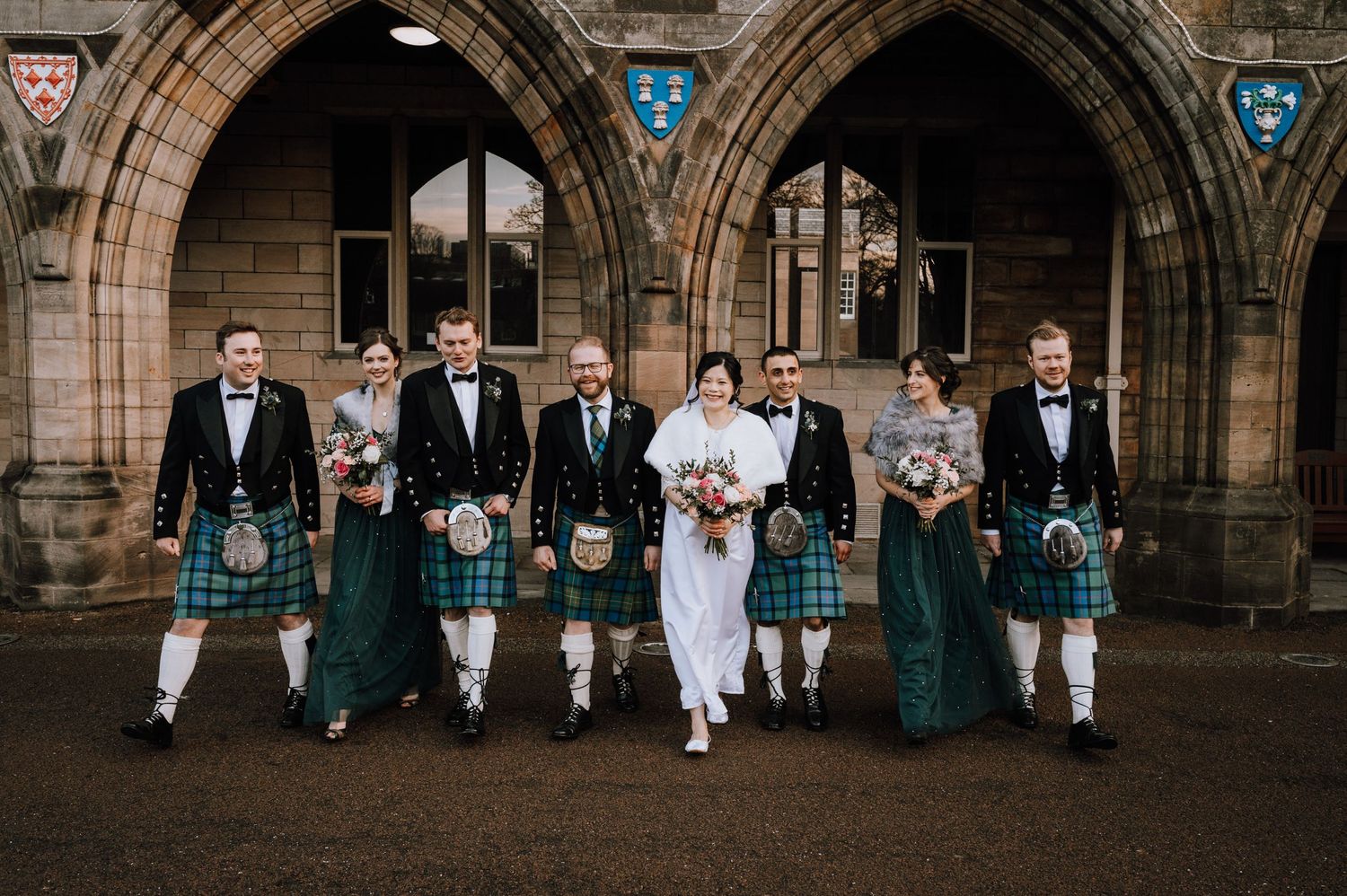 A wedding party in Scottish formal wear walks beneath Gothic church arches.