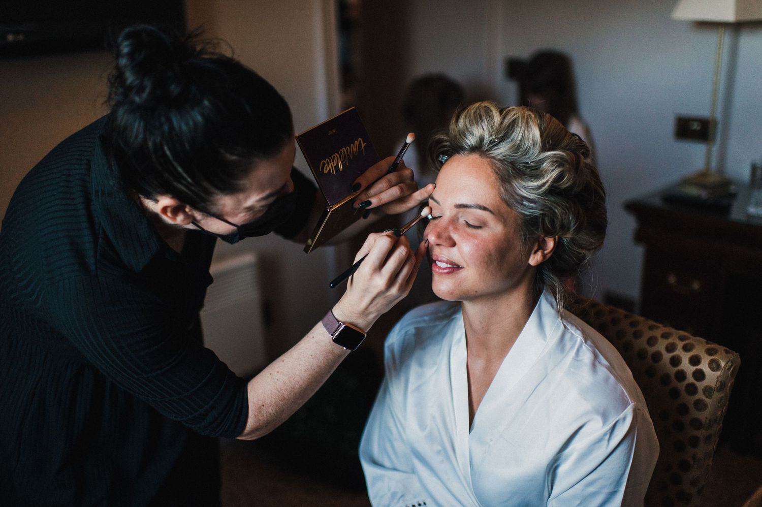Makeup artist applying cosmetics to client in white robe during preparation session.