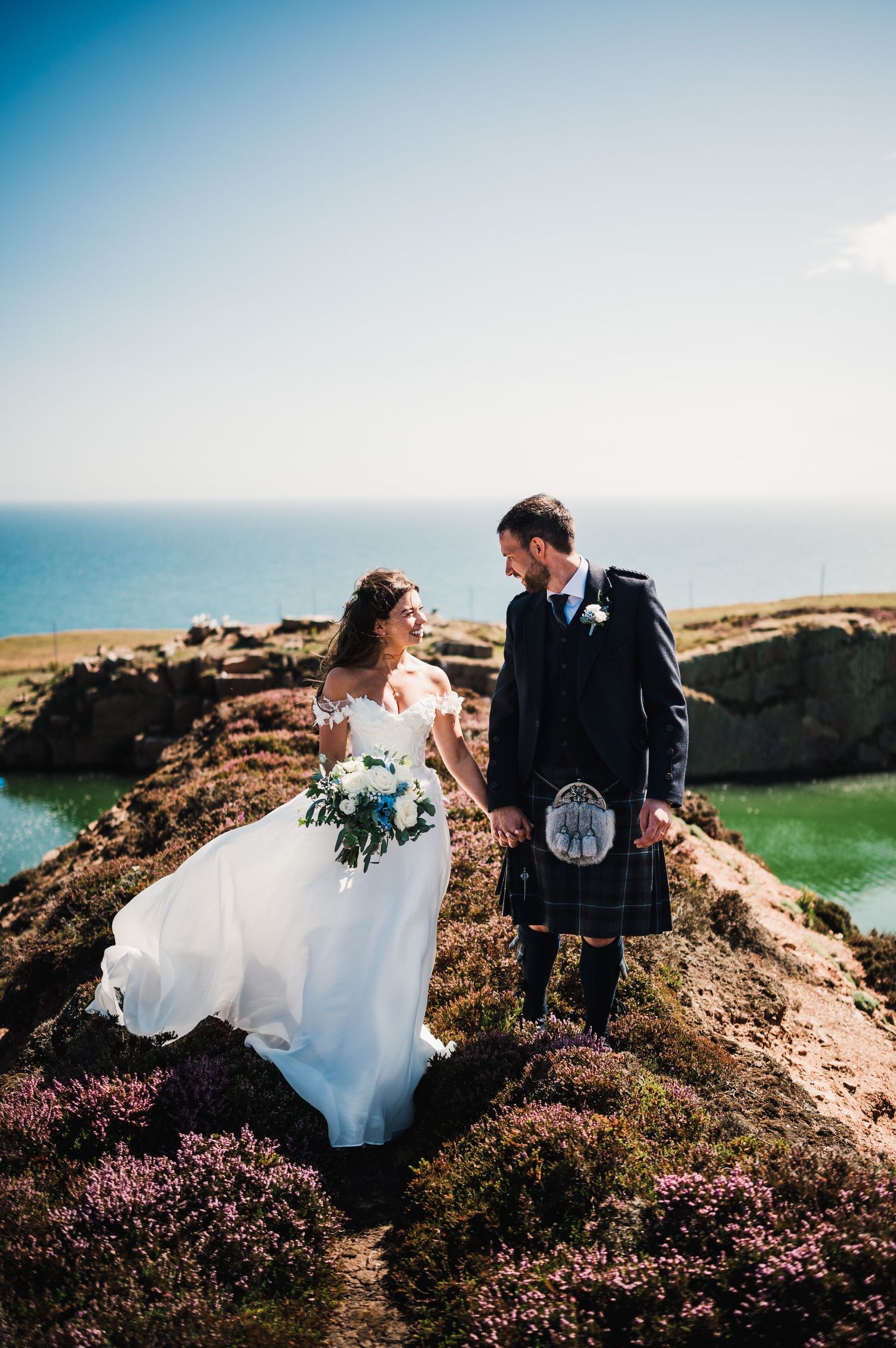 Bride in flowing dress and groom in traditional kilt pose on dramatic coastal clifftop.