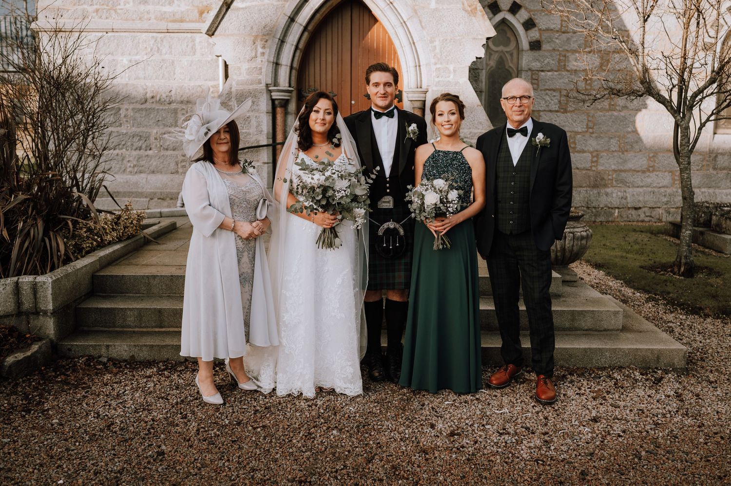 Wedding party in mix of formal wear and Scottish attire pose on church steps.