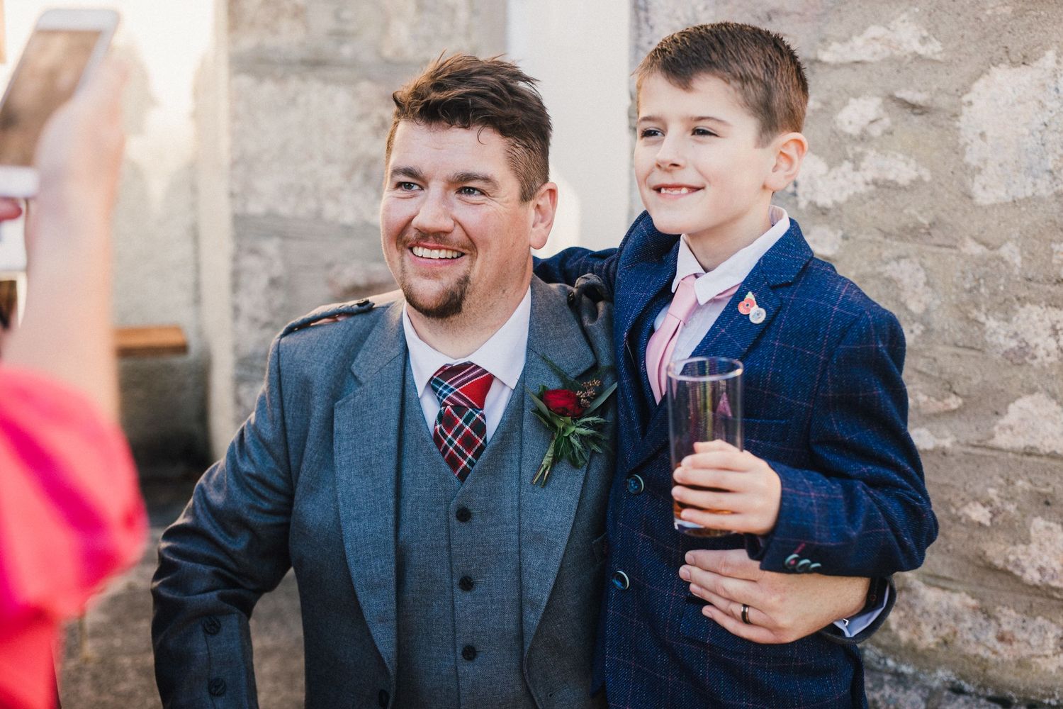 A heartwarming moment between wedding guests in formal attire against a stone wall.