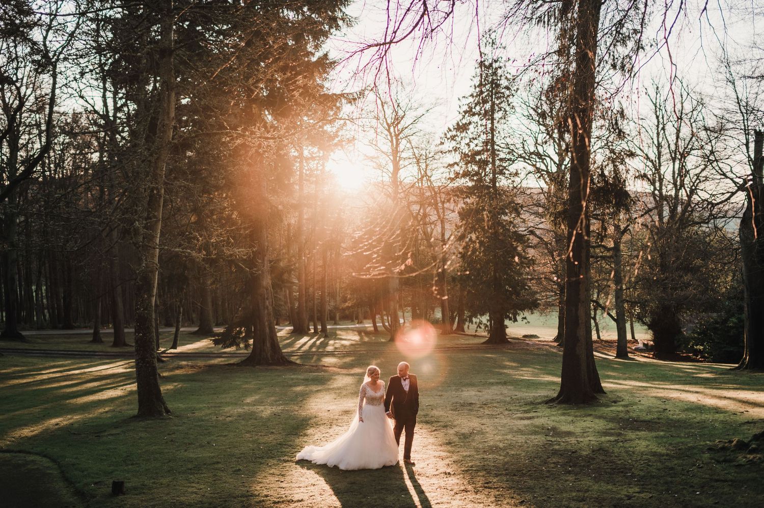 Sunset wedding photo captures couple walking through tree-lined path with golden light streaming through.