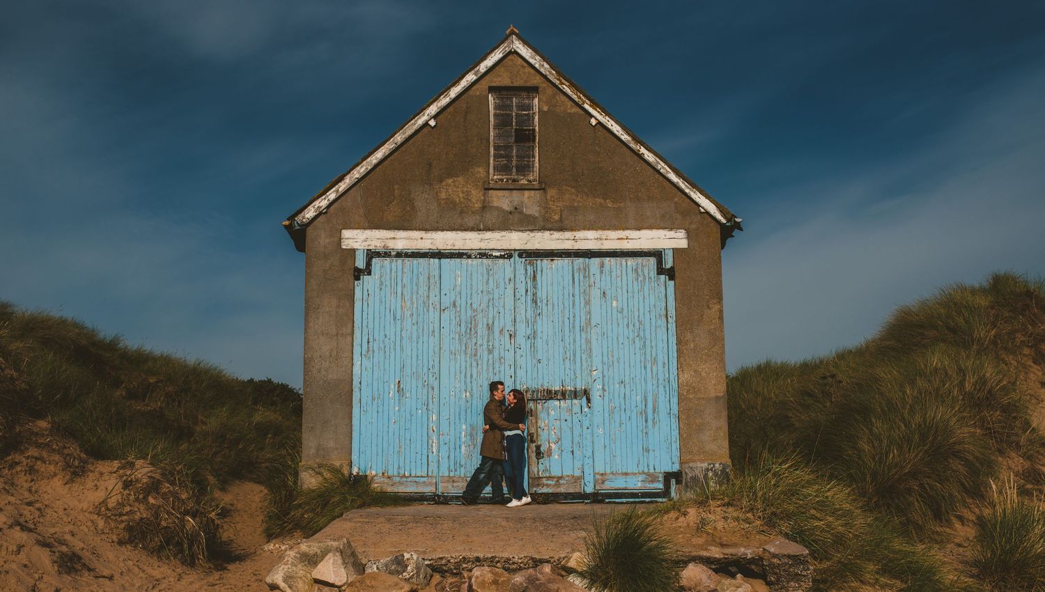 A rustic blue-doored barn standing against a moody sky on coastal grounds.