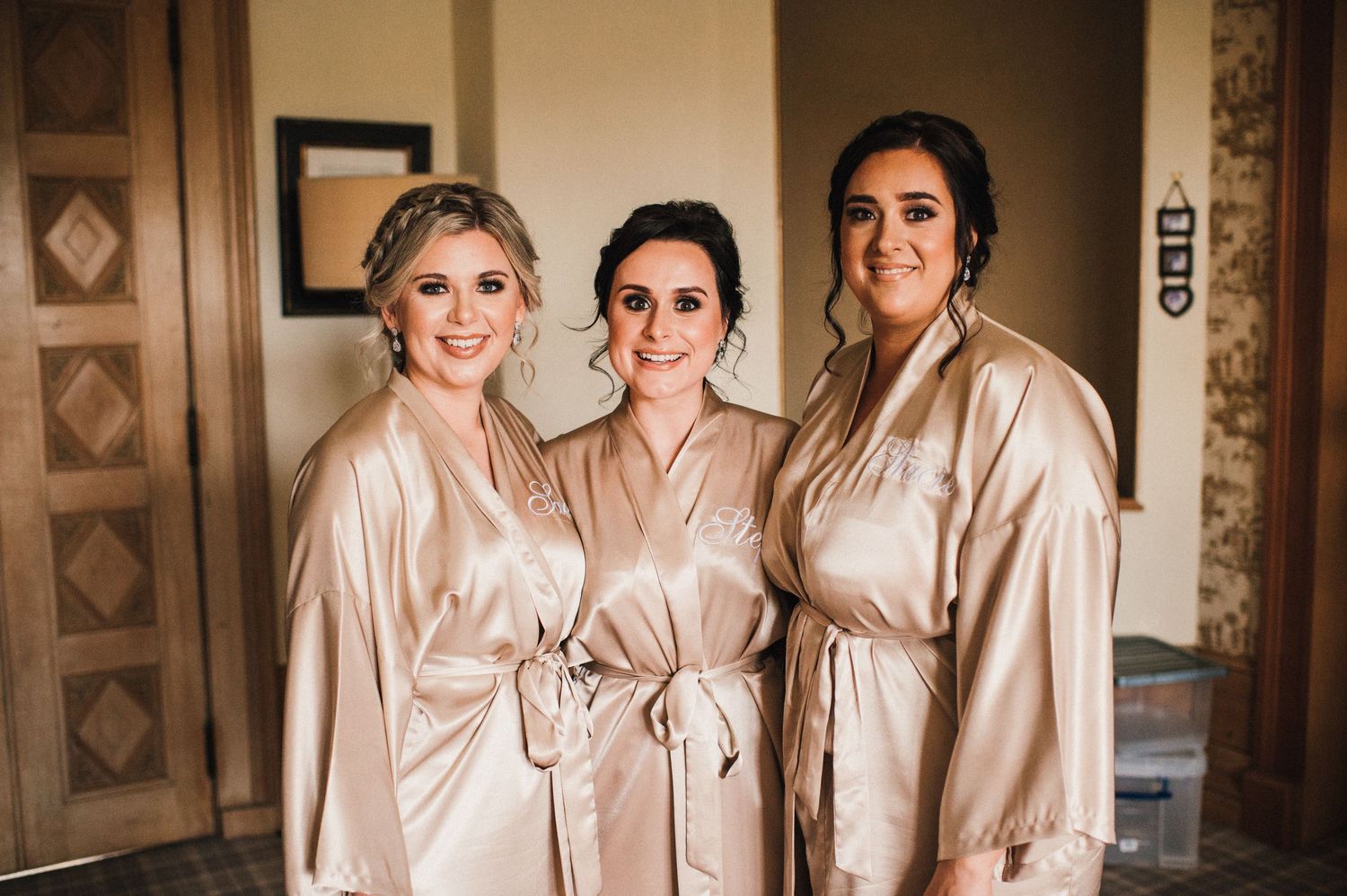 Three bridesmaids pose together wearing matching champagne-colored silk robes during wedding preparations.