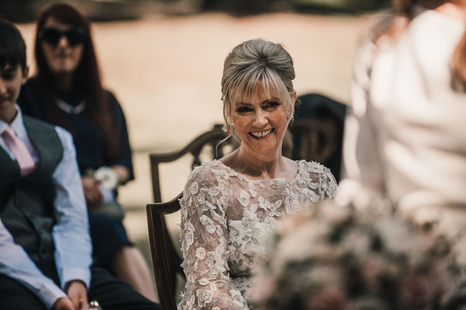 Guest in elegant lace dress smiles during outdoor wedding celebration.