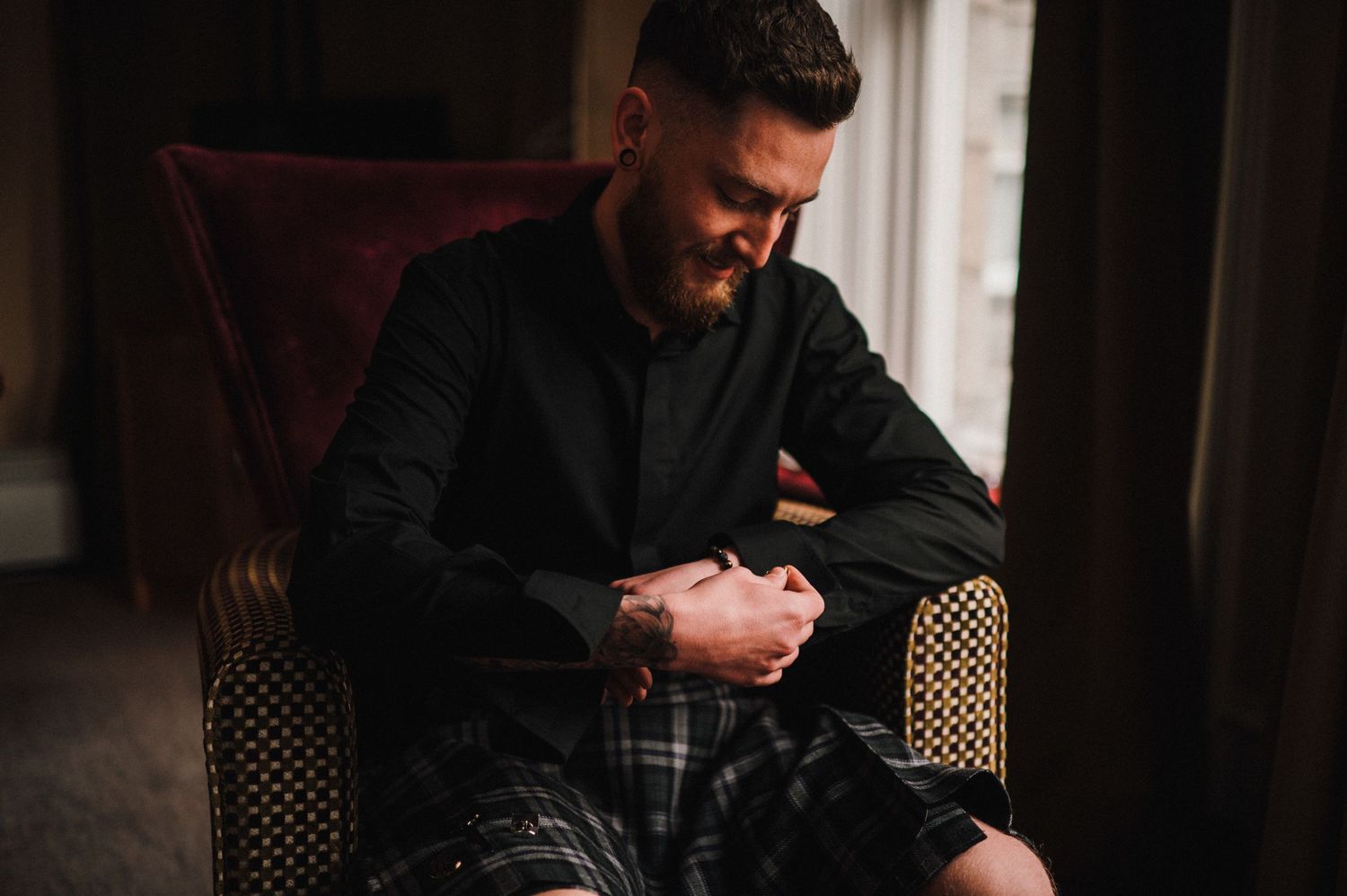 Person in black attire and plaid kilt seated in an elegant armchair reading correspondence.