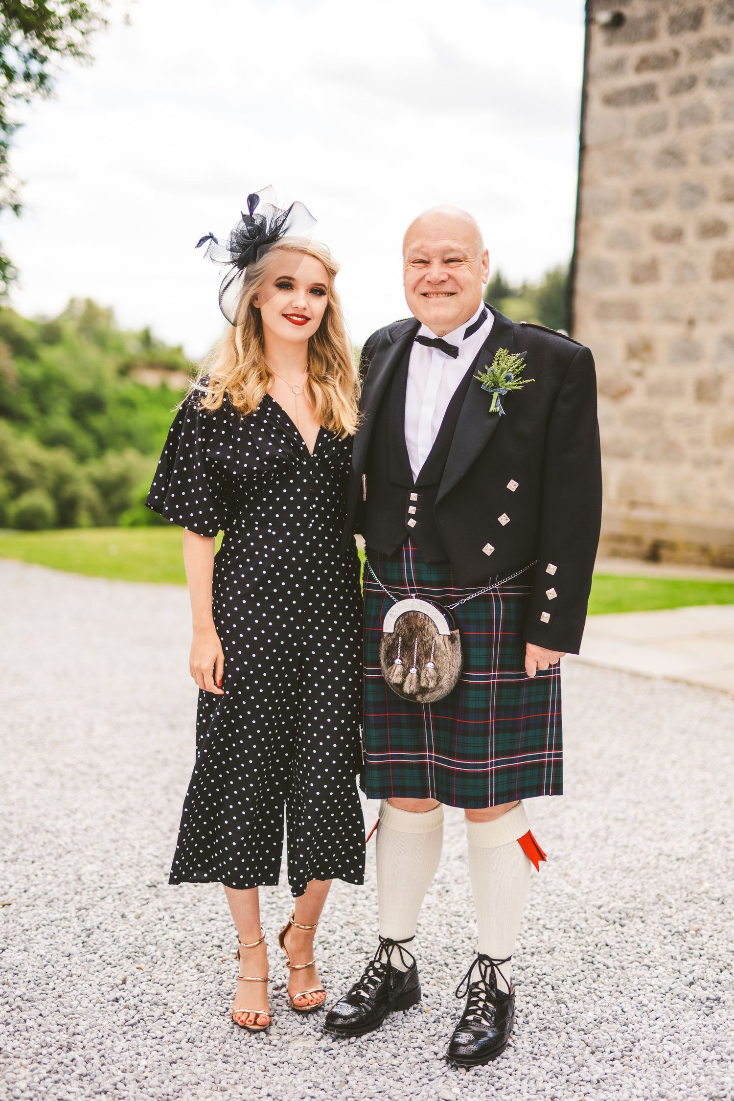 A polka dot dress and traditional Scottish kilt are worn by a couple posing on a gravel path.