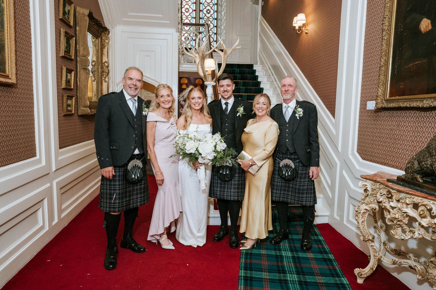 Wedding party members posing on grand staircase in traditional Scottish attire.