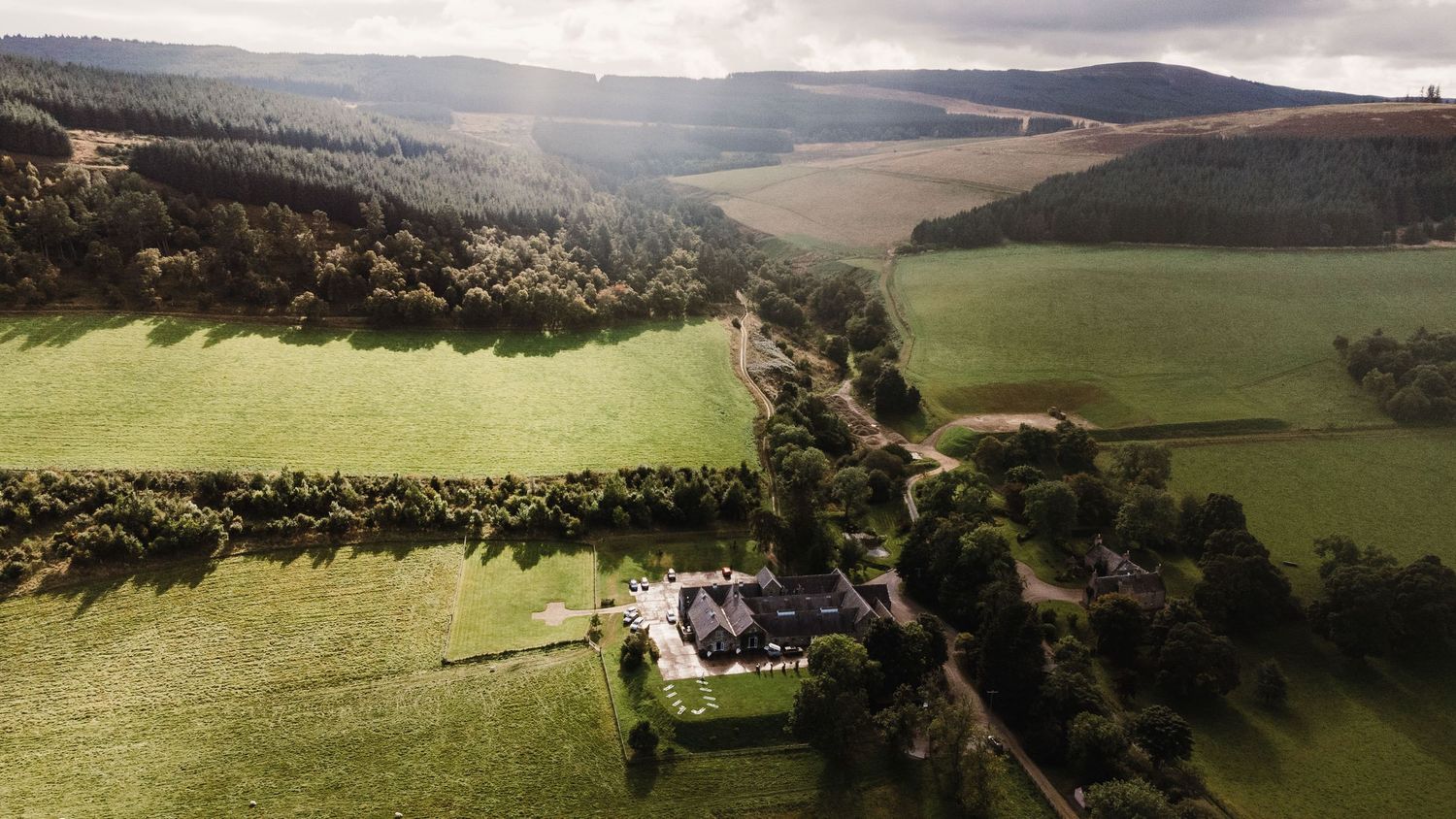 Panoramic aerial view of rolling green hills and farmland with mountains in the distance under dramatic cloudy skies.