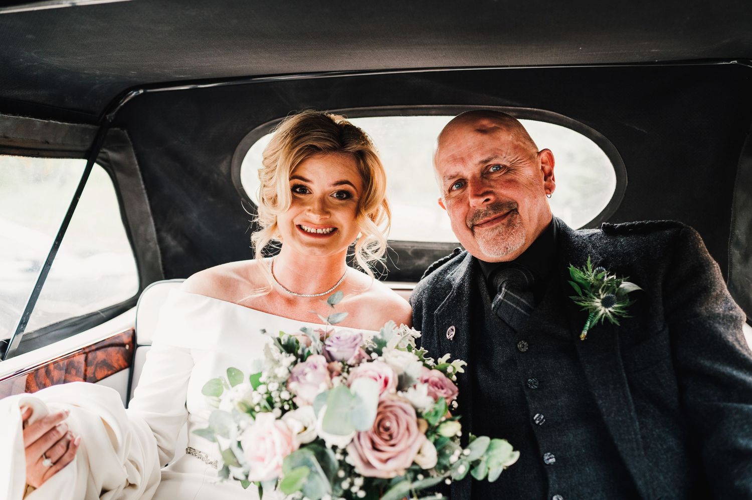 A black and white wedding photograph showing a couple in a car with bouquets and smiles.