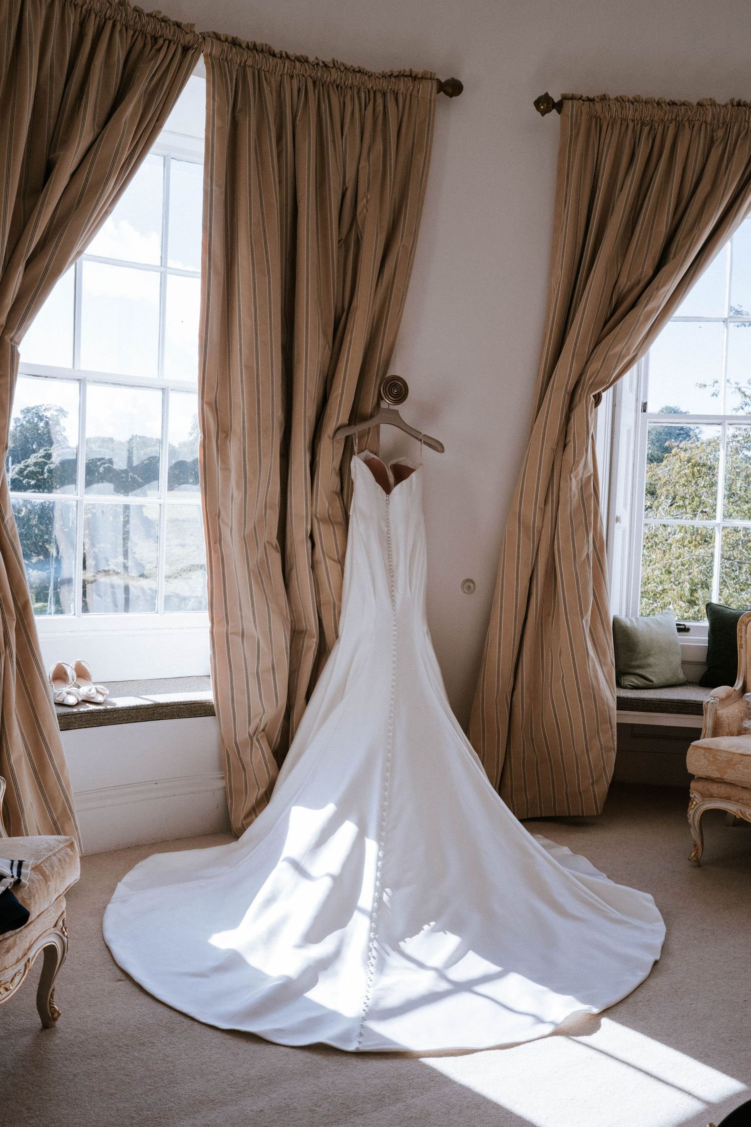 White wedding dress displayed in sunlit room with elegant taupe curtains and period windows.