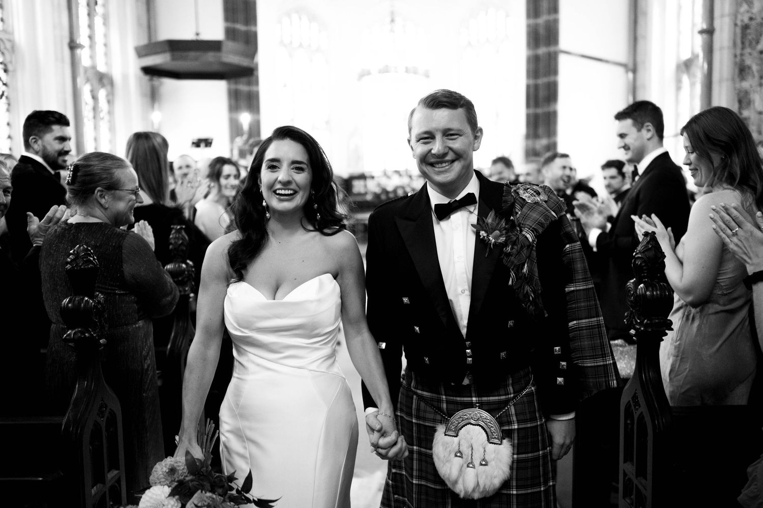 Black and white photo of newlyweds walking down church aisle in Scottish wedding attire.