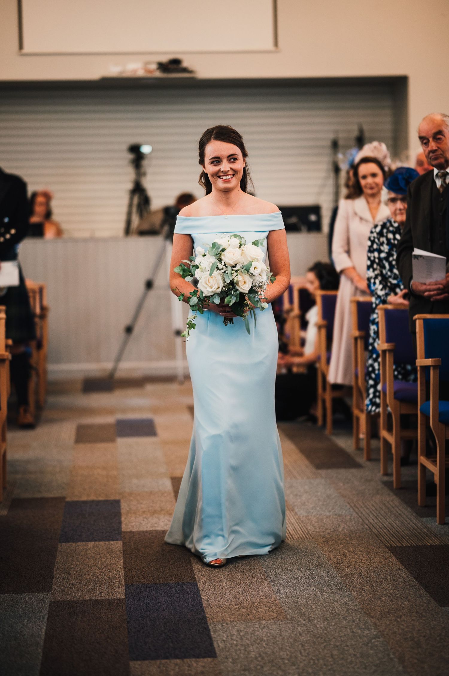 Bridesmaid walks down church aisle wearing light blue off-shoulder dress carrying white floral bouquet.