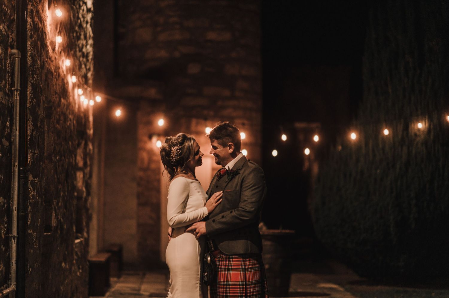 A romantic moment between newlyweds under string lights against a rustic brick wall.