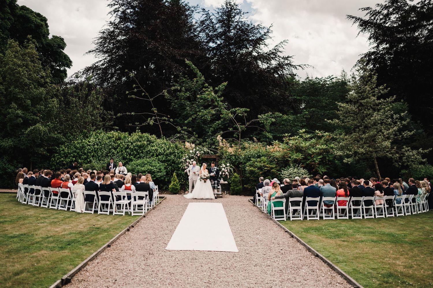 A beautiful outdoor garden wedding ceremony with white chairs arranged in rows along a gravel path.