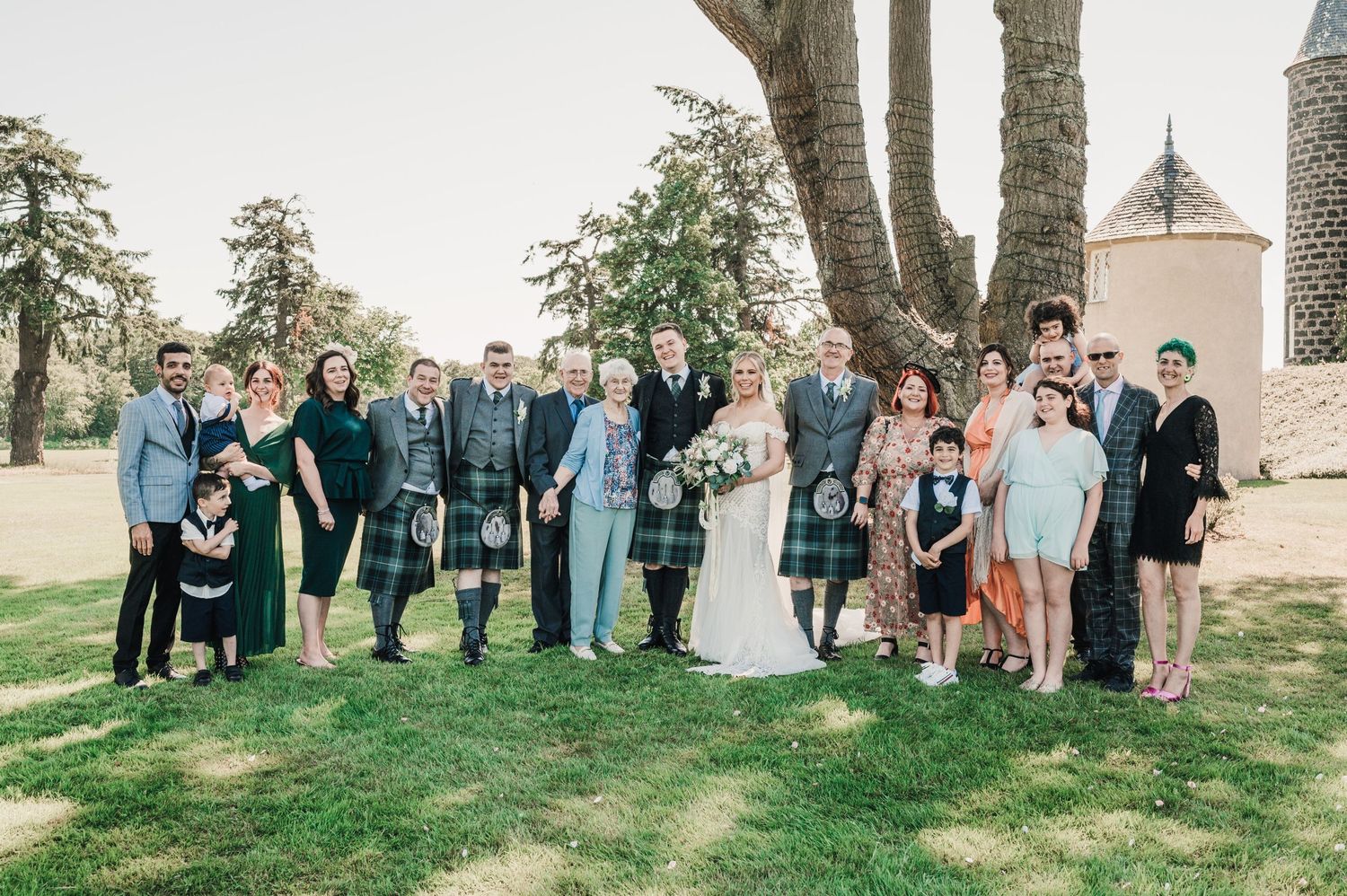 Wedding party gathered on green lawn in front of stone building with mix of formal attire and kilts.