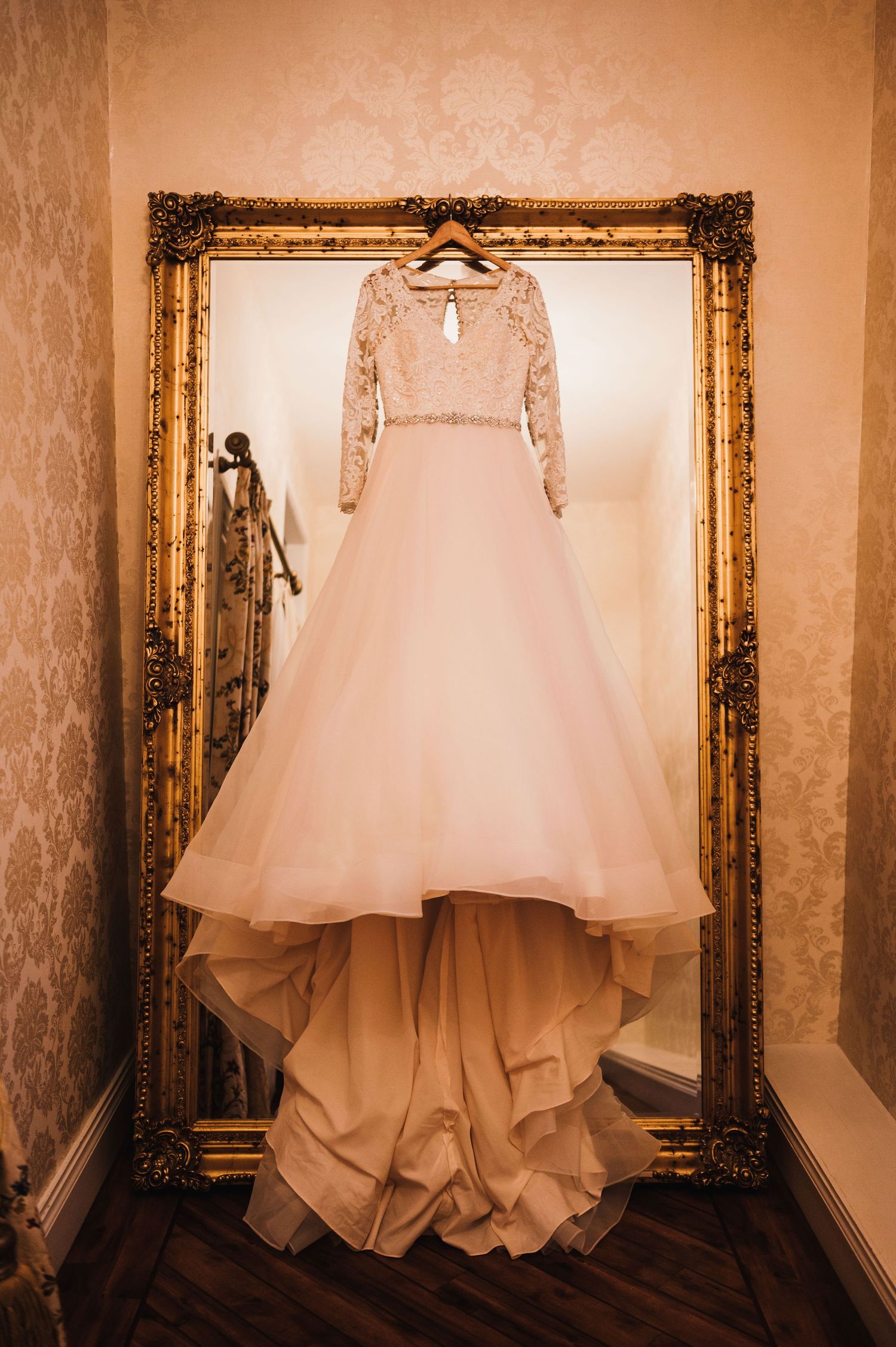 Elegant wedding dress with lace sleeves displayed on hanger in ornate gold mirror frame.
