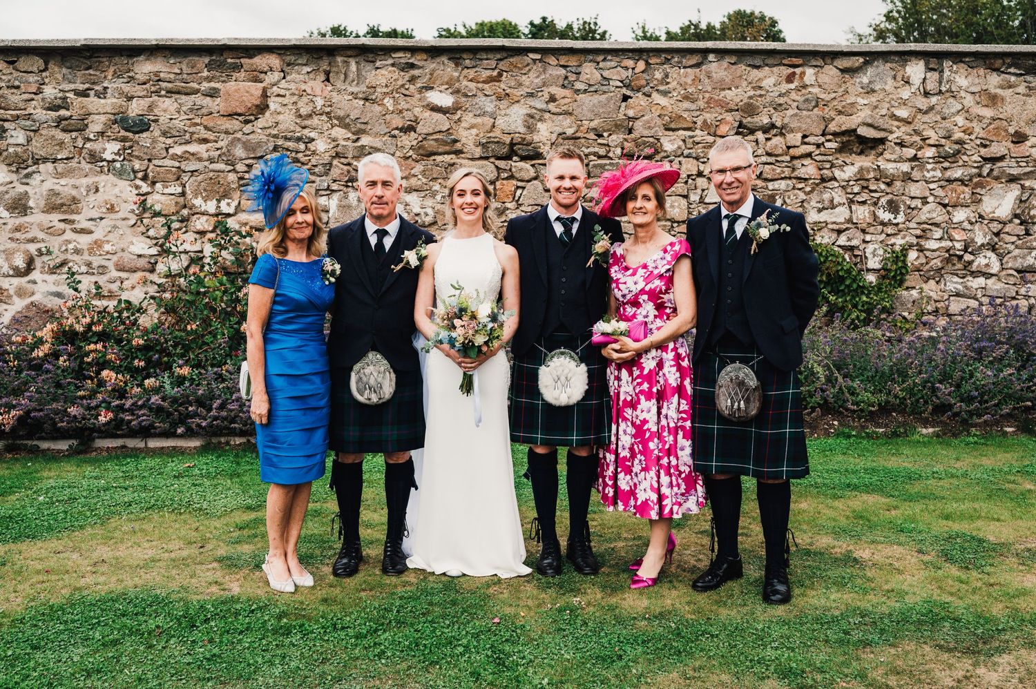 Wedding party wearing traditional Scottish kilts poses against stone wall backdrop at outdoor ceremony.