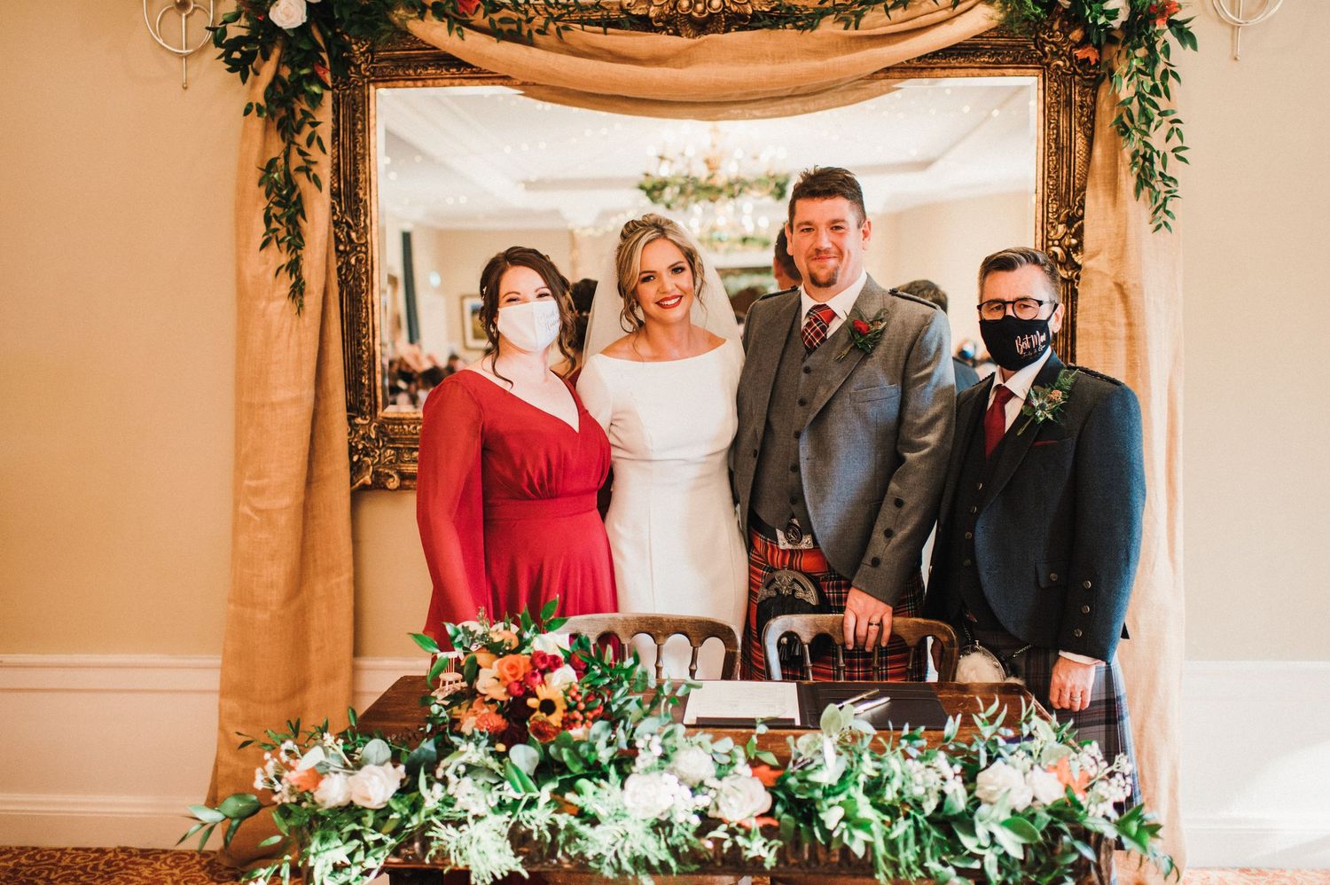 Wedding party poses under floral arch with burgundy and green decor at ceremony.