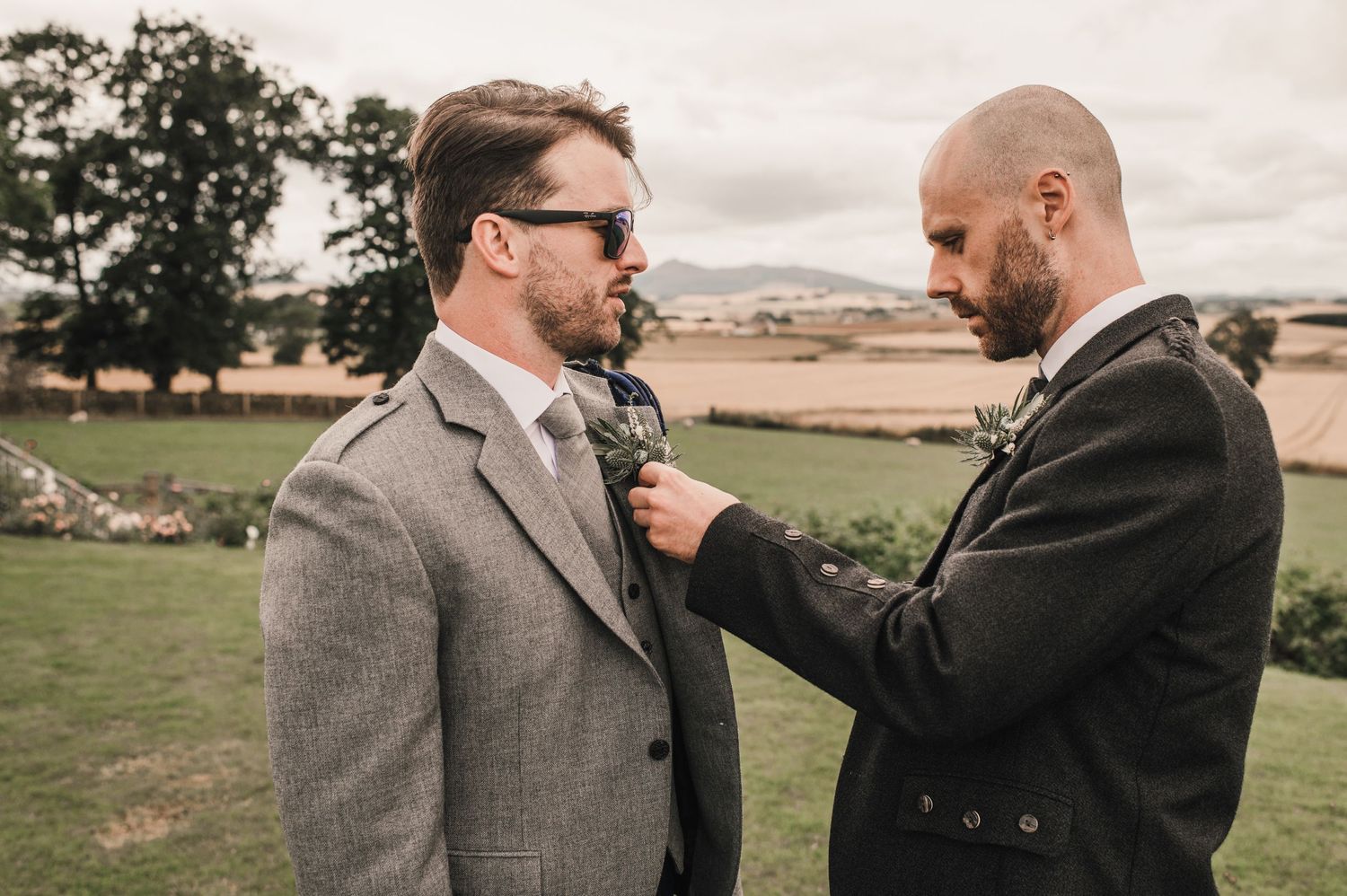 Two wedding attendants helping each other with boutonnieres against rolling countryside backdrop.