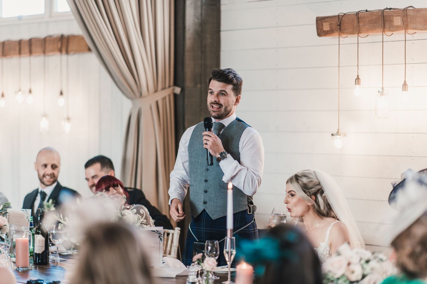 A speaker gives a toast at an elegant wedding reception with hanging pendant lights and white brick walls.