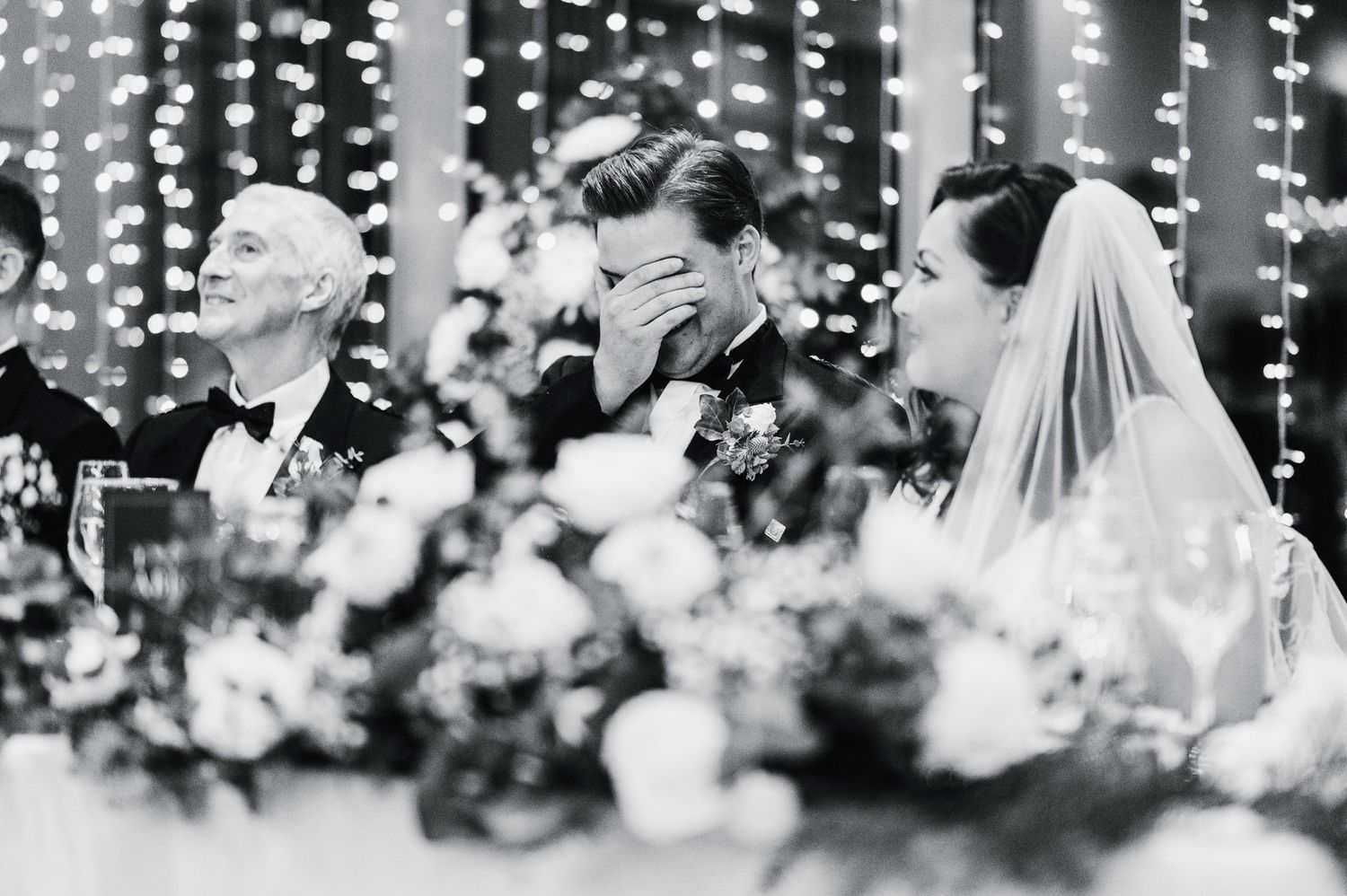 Black and white shot of emotional wedding reception moment with string lights and floral decor.
