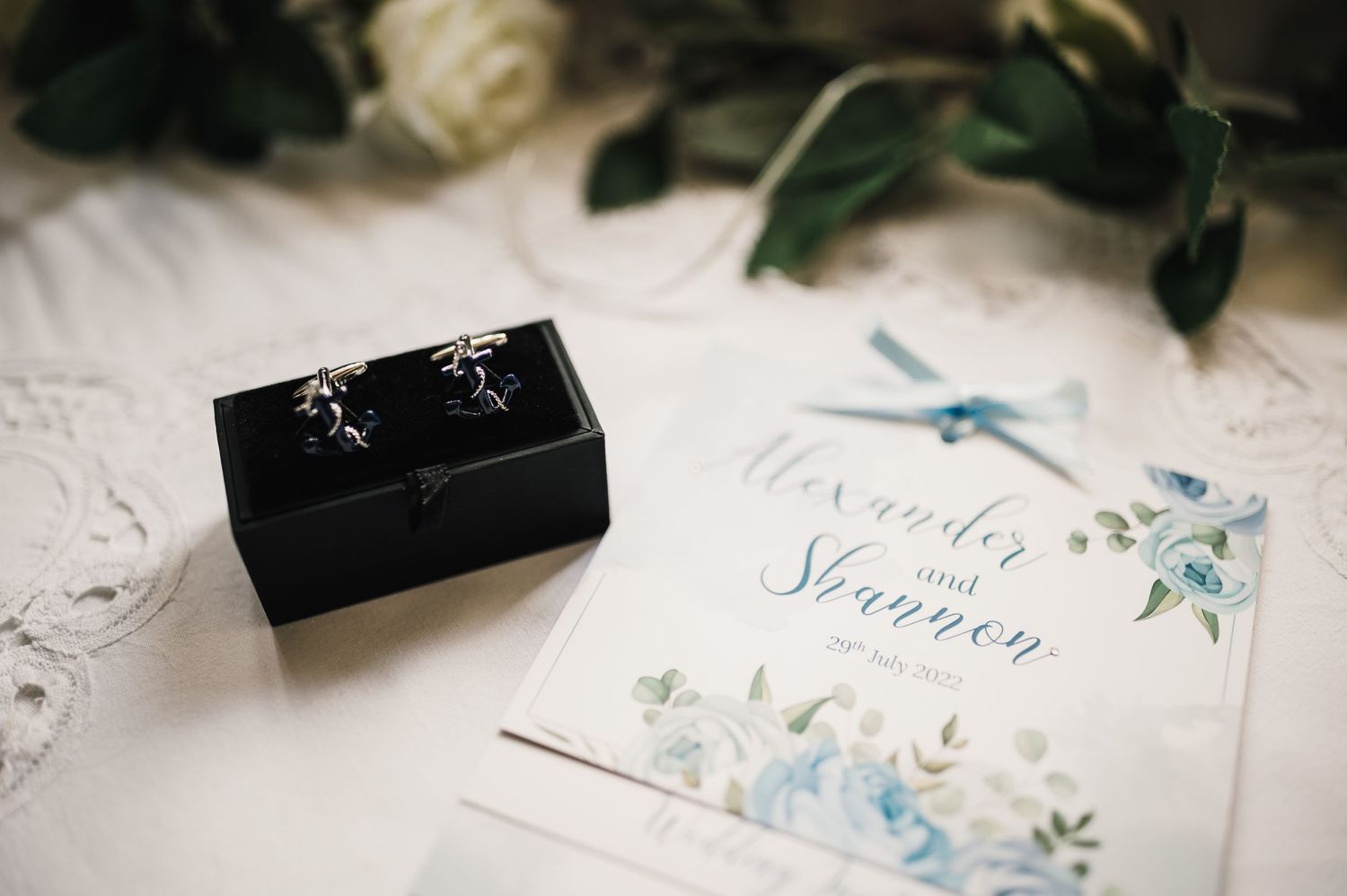 Wedding invitation with blue floral design next to black cufflink box on white surface.
