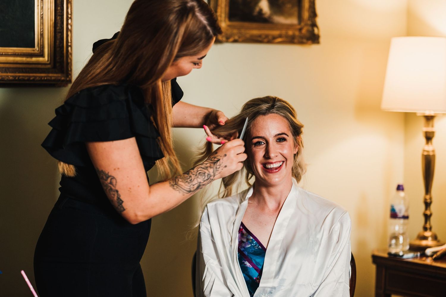 A makeup artist applies cosmetics to someone wearing a white robe in an elegant room with warm lighting.