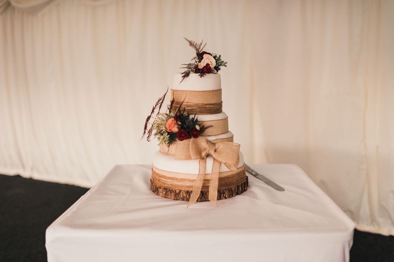 A rustic wedding cake with floral decorations sits on a wooden slice against a white backdrop.