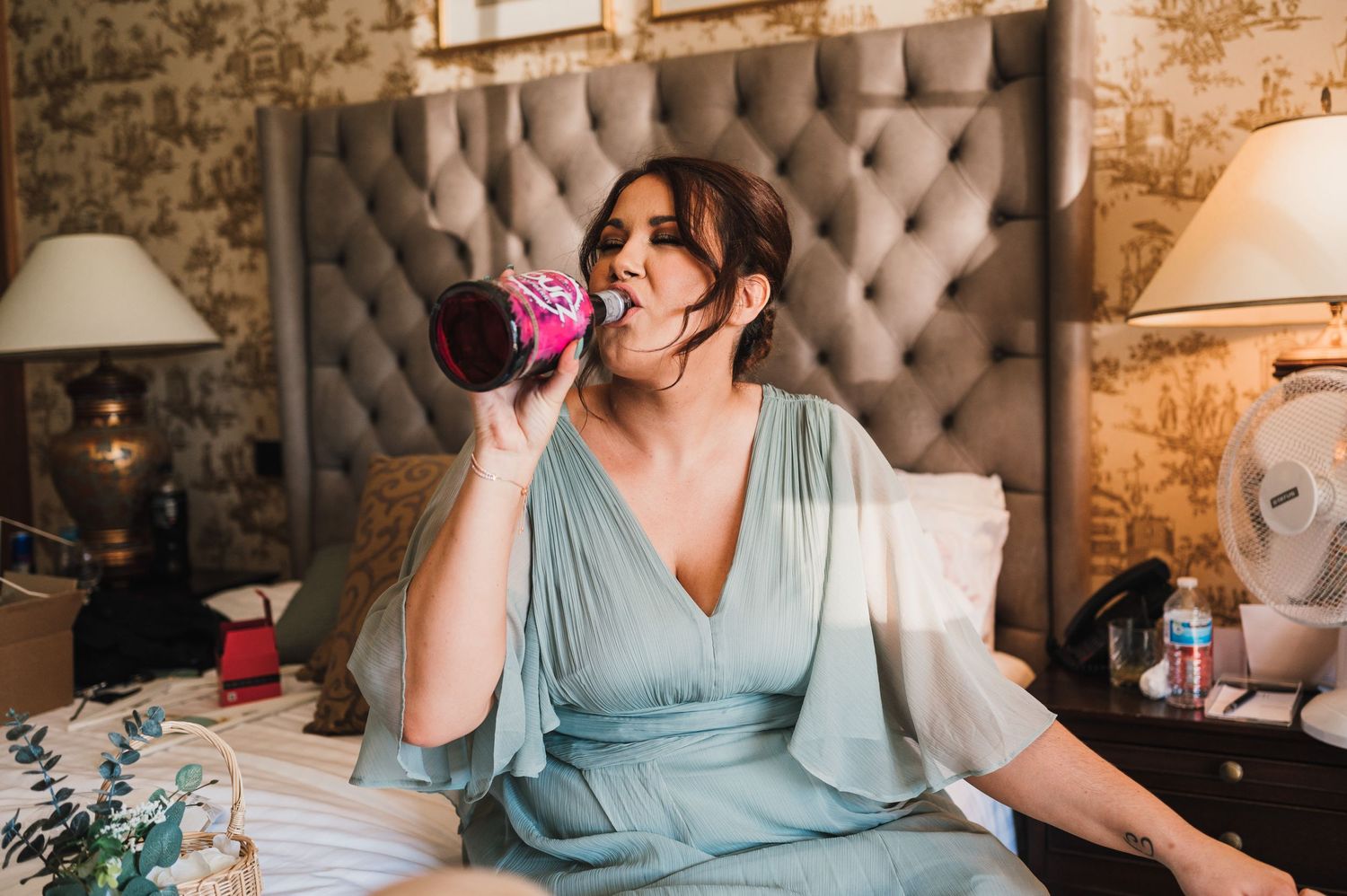 Someone sits on a tufted headboard bed while drinking from a pink beverage can.