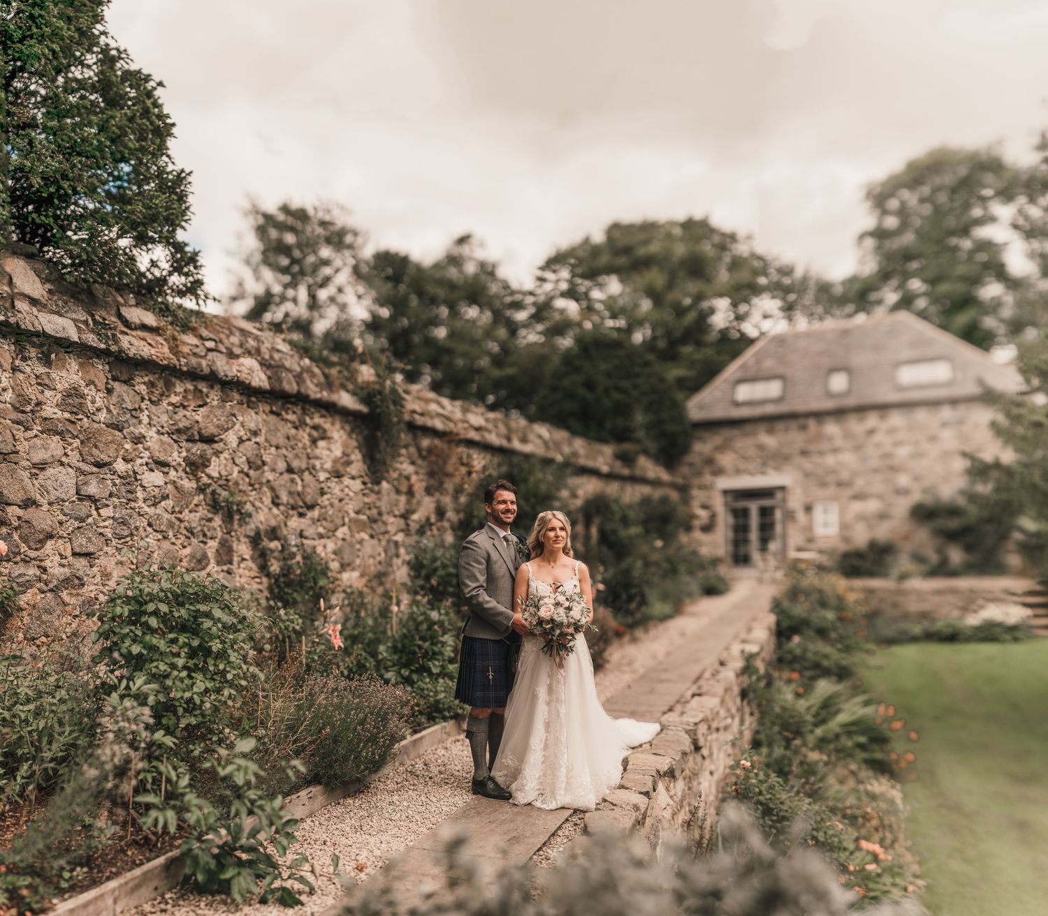 Couple poses on stone wall near historic castle building during Scottish wedding celebration.