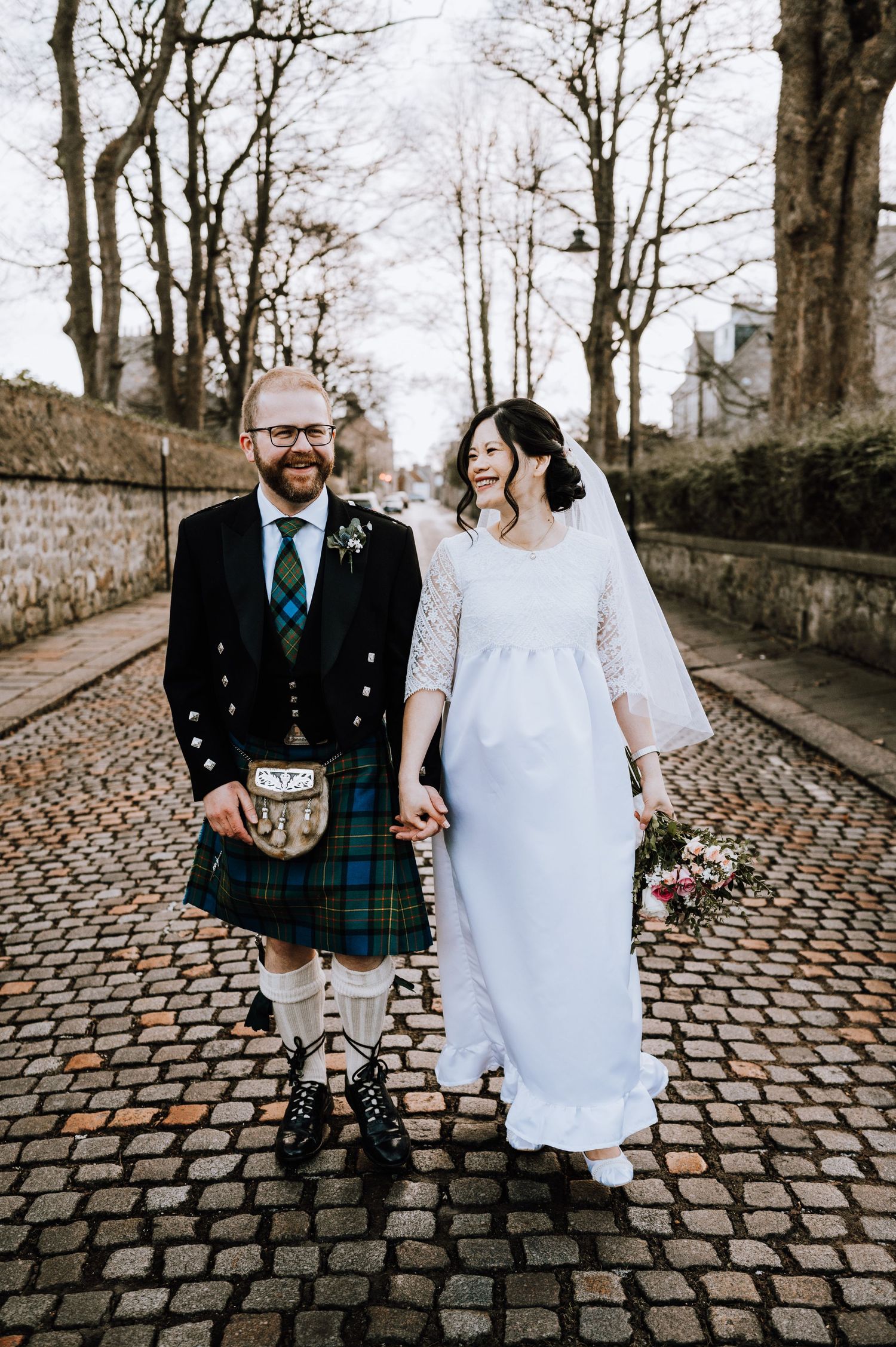 A couple in Scottish wedding attire stands together on a historic cobblestone street.