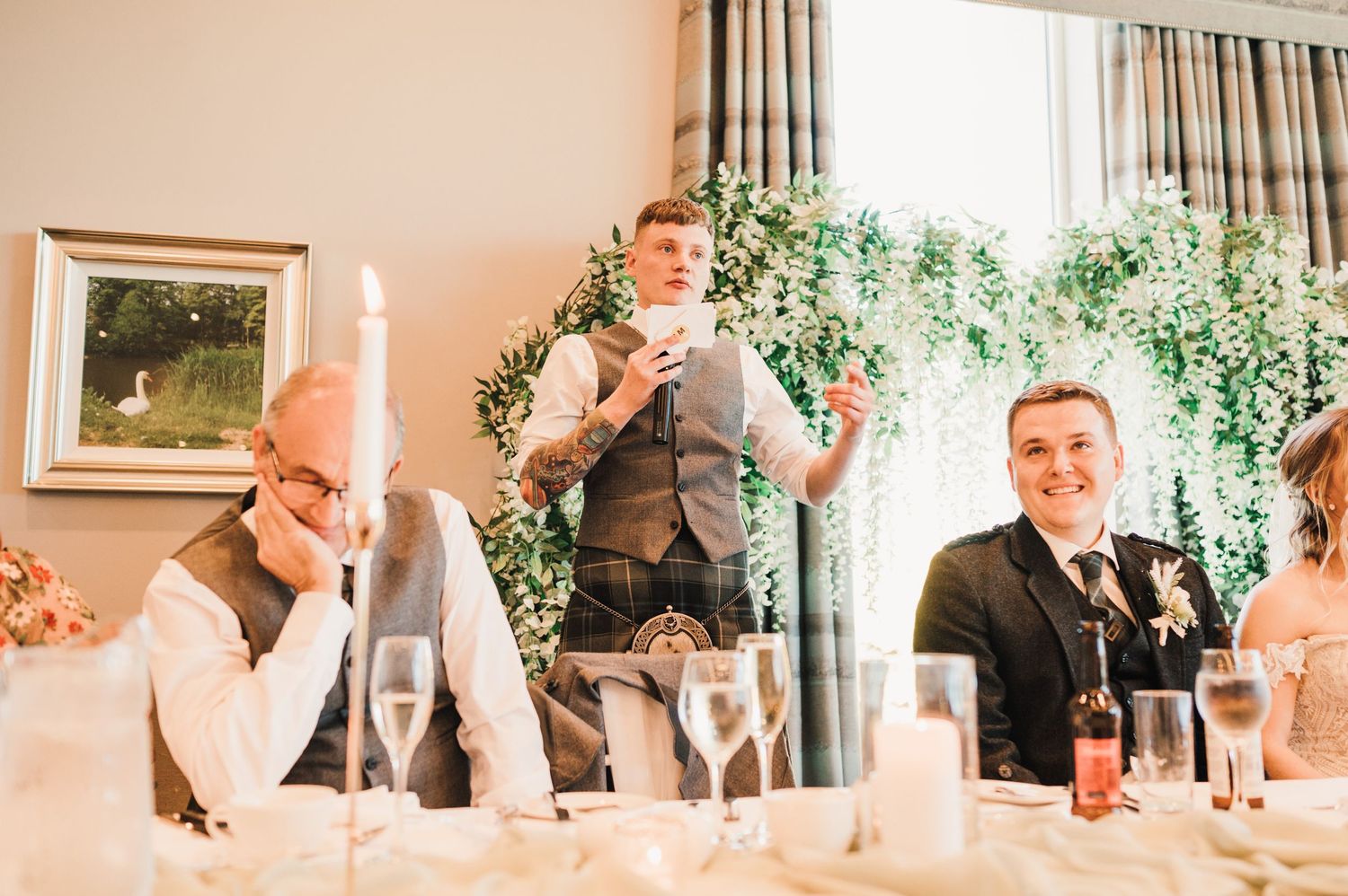 Guests laugh during speeches at a wedding reception with floral decor and champagne glasses.
