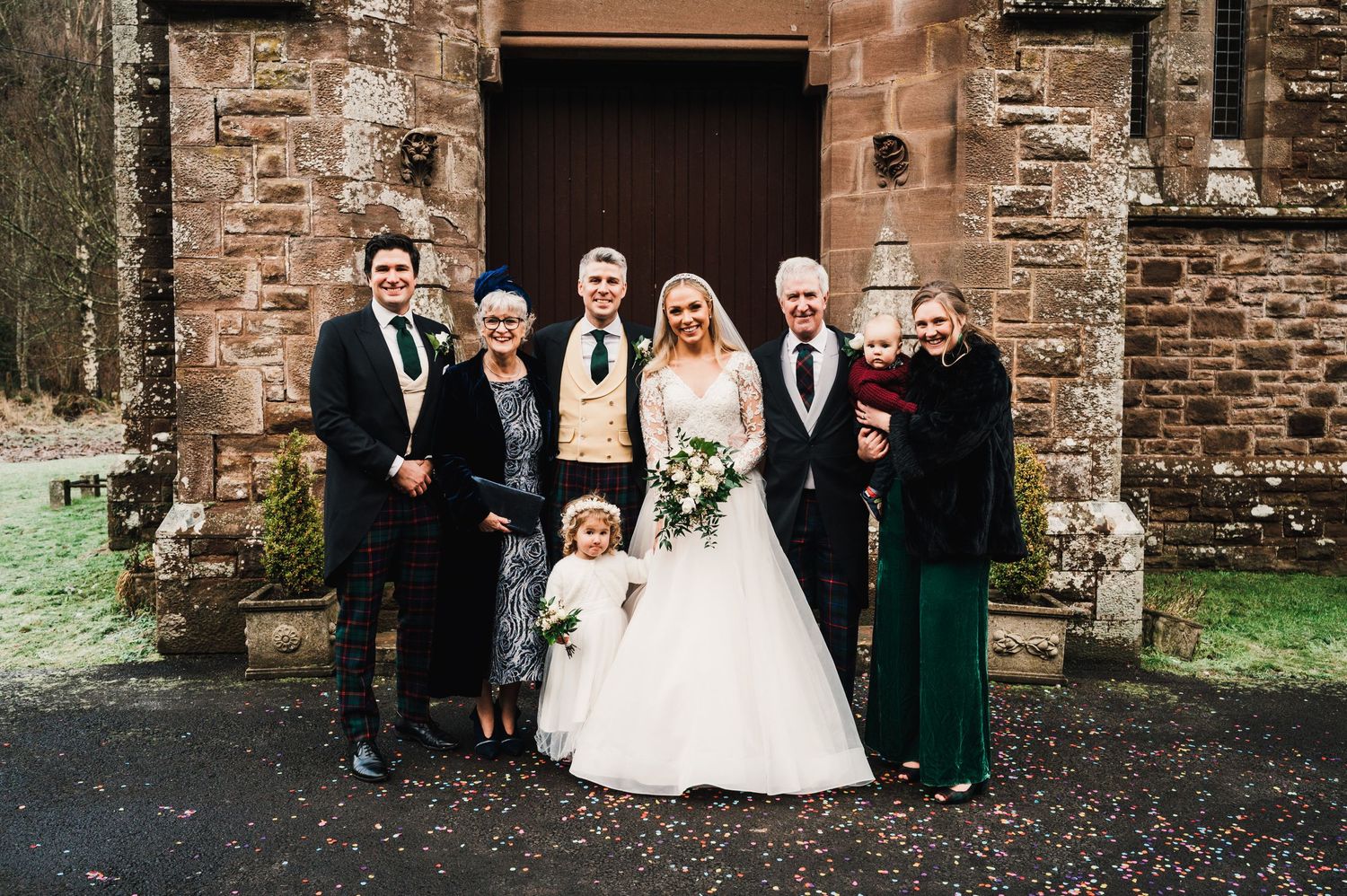 Small wedding group gathers for portrait outside stone church entrance in formal attire.