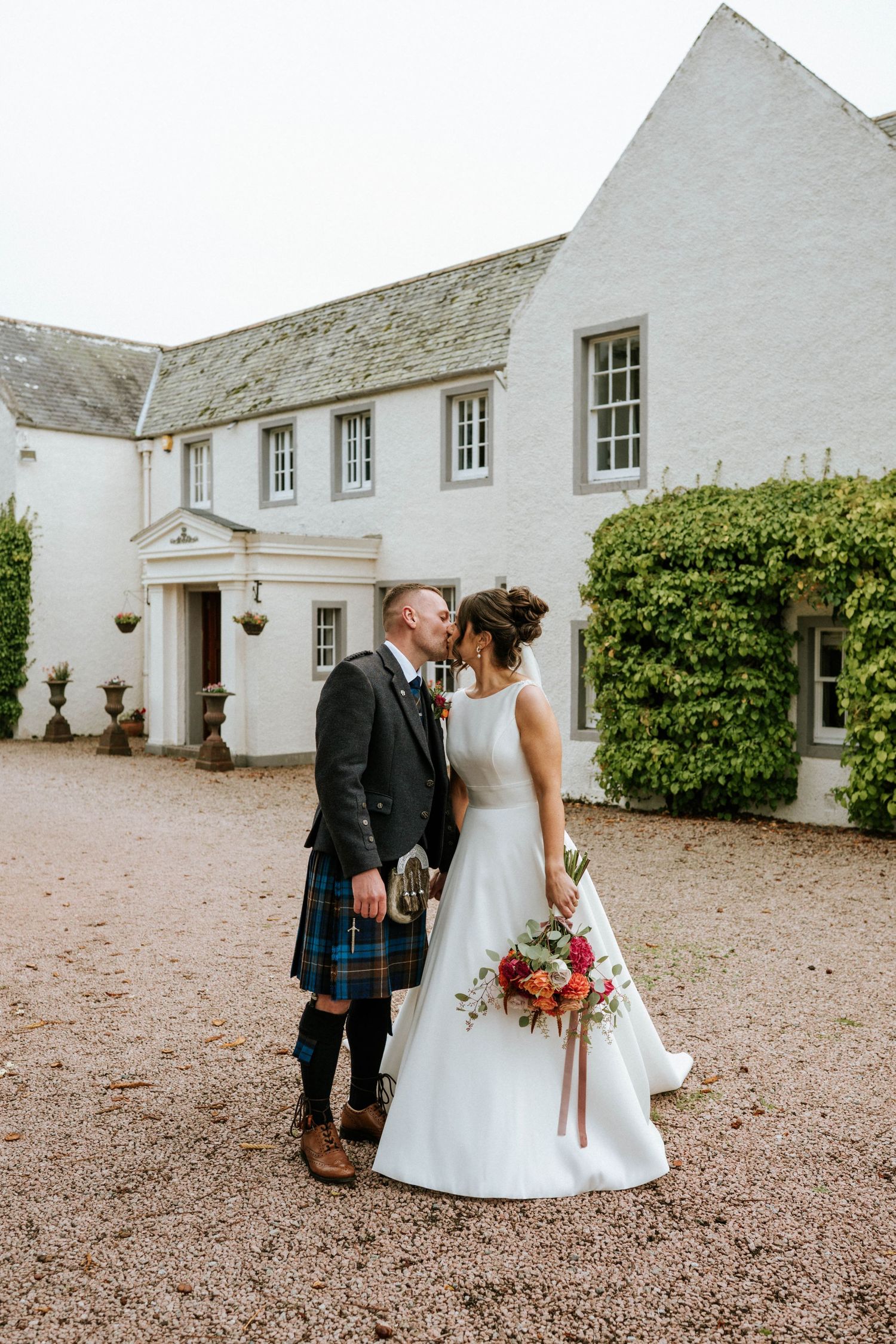 A couple stands in front of a white colonial-style house during their Scottish-themed wedding.