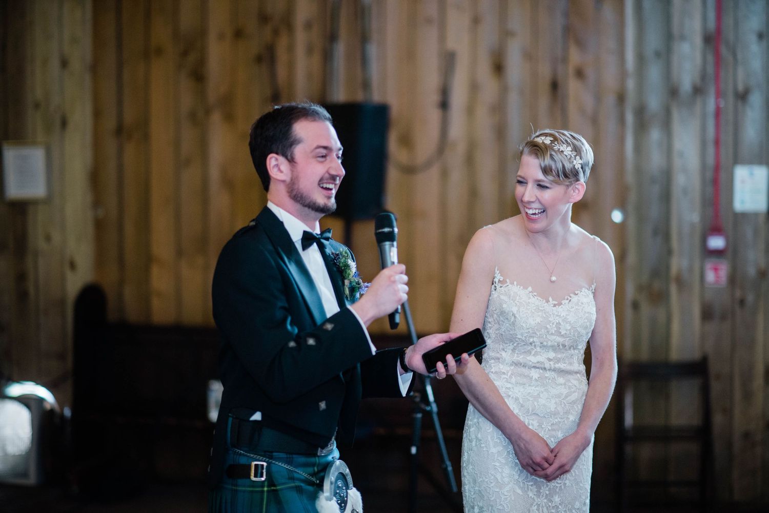 Couple sharing laughs during wedding reception speeches in wood-paneled room.