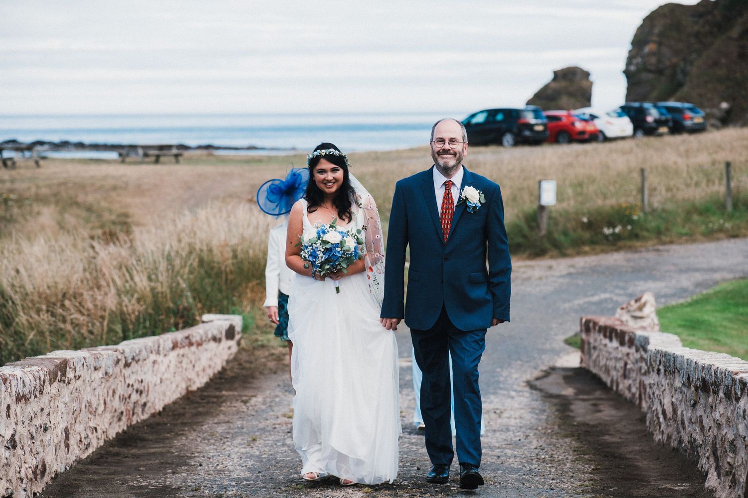 Couple walks along stone-walled pathway near coastal cliffs.