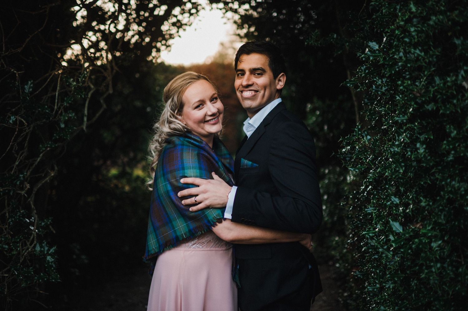 A couple embraces on a nature path at sunset wearing formal wear and a plaid shawl.