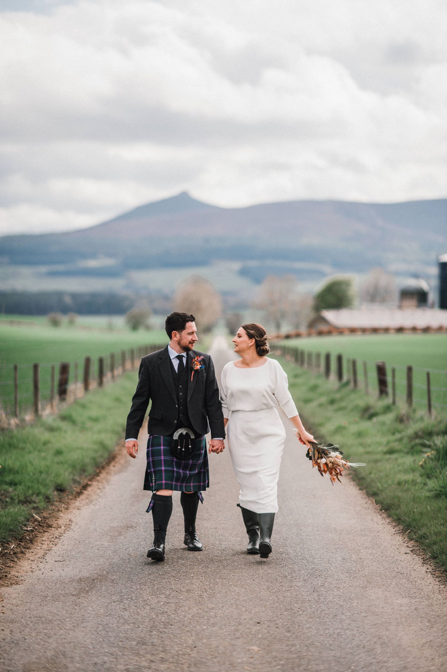 Scottish wedding couple walking hand in hand down a rural path with mountains and farmland behind them.